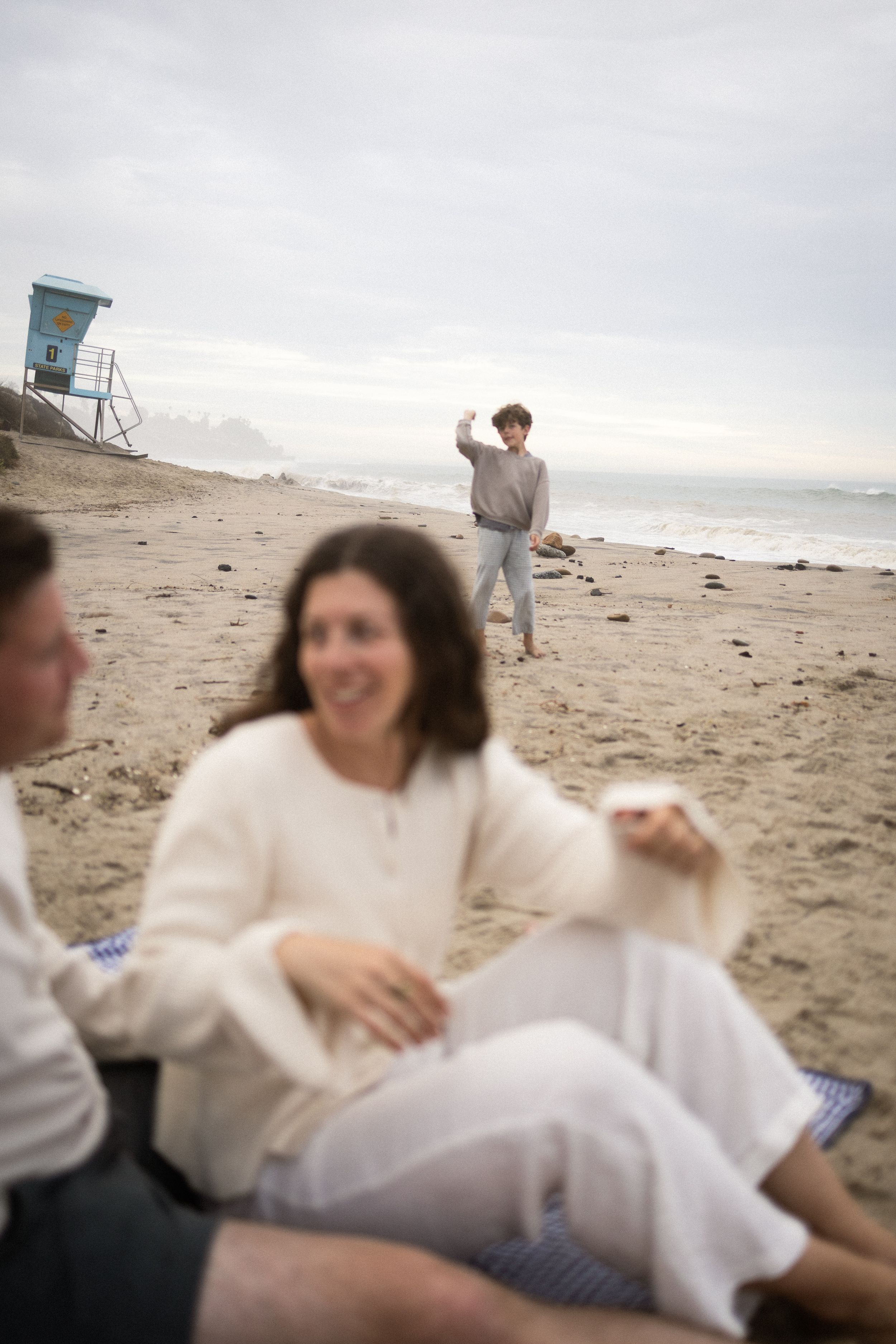 Friends relaxing on a sandy beach near a lifeguard tower on a cloudy day, with a person waving in the background.