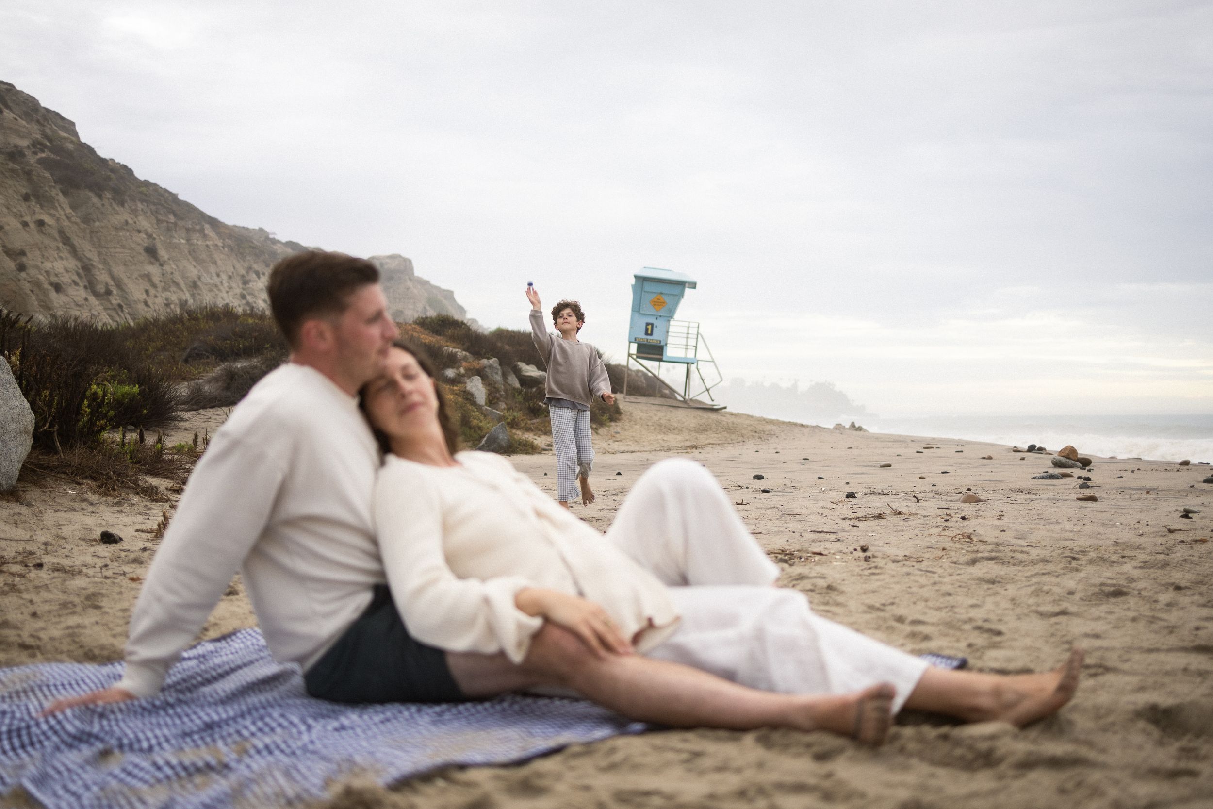 Couple relaxing on beach blanket while child plays near lifeguard tower on sandy California coastline.