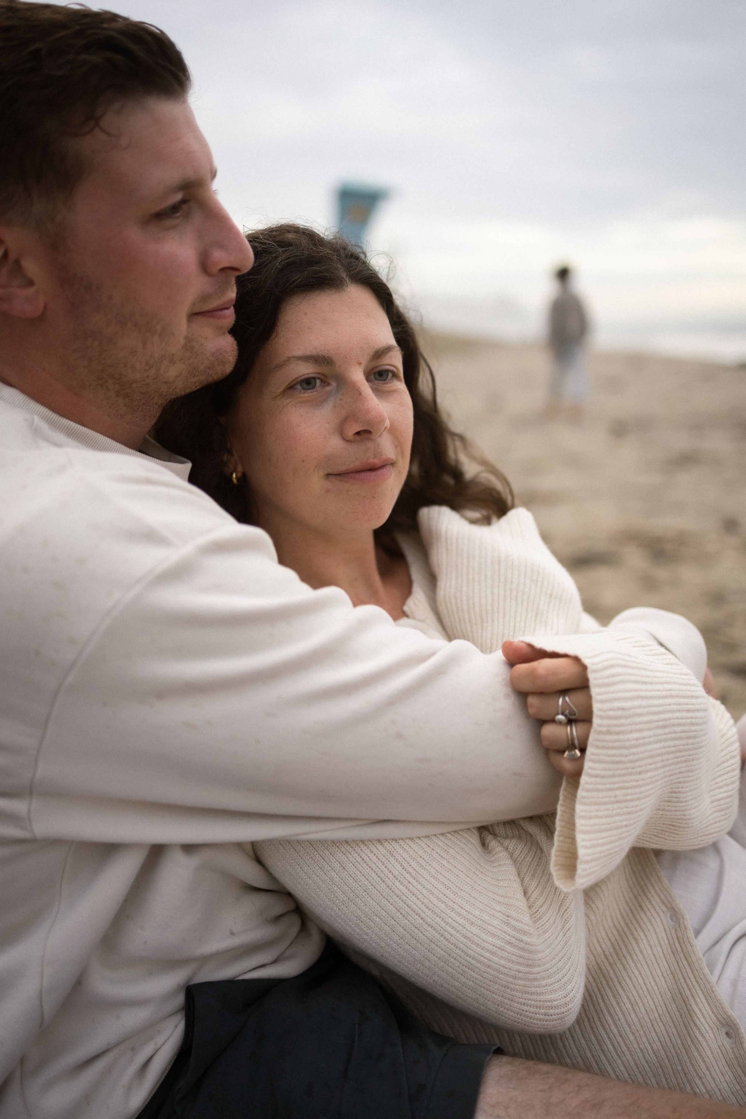 Couple embracing on a cloudy beach, woman in white knit sweater with rings, man hugging her from behind.