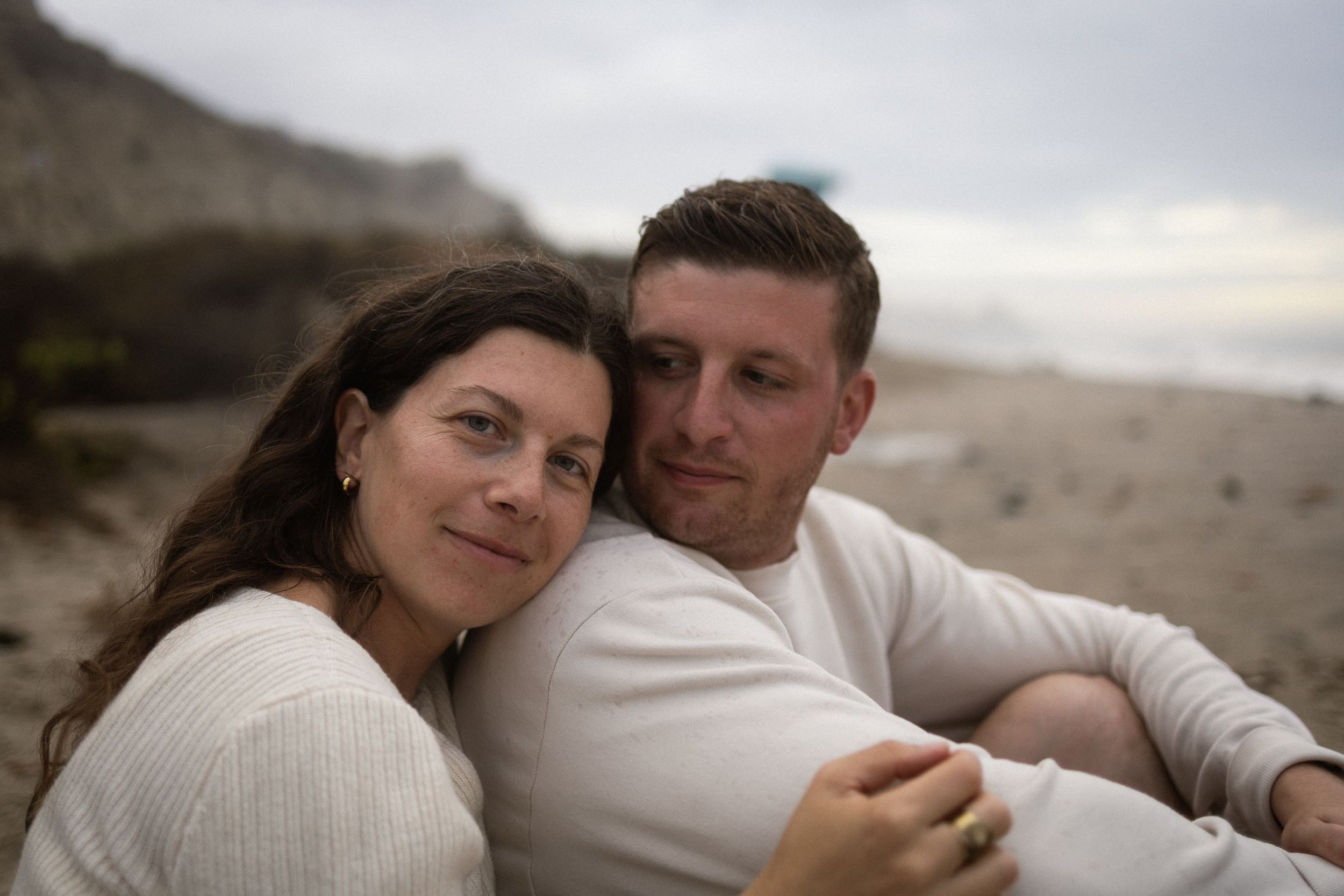 A couple sits closely together on a sandy beach, smiling warmly, dressed in white tops with cliffs in the background.