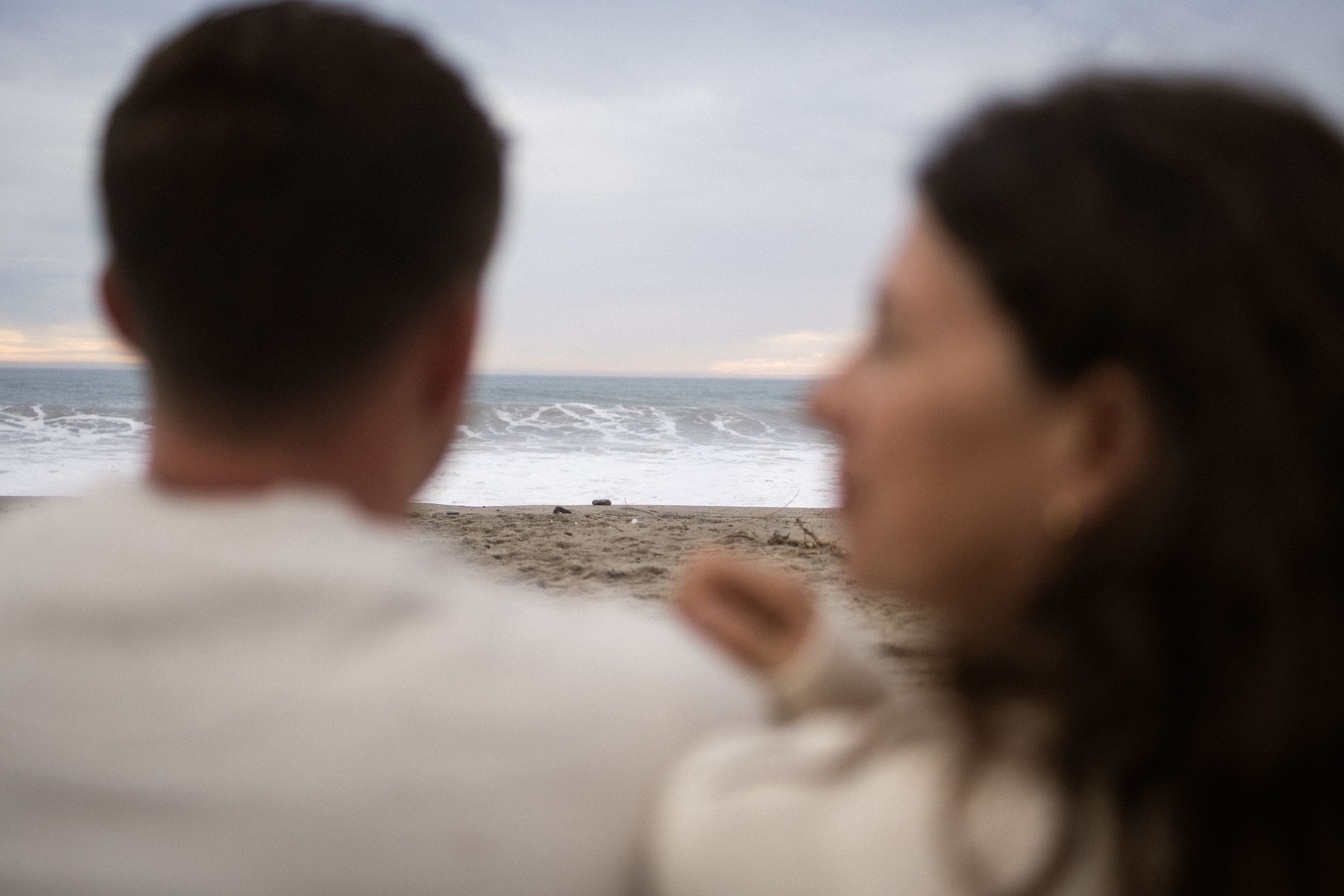 Couple seen from behind gazing at ocean waves on a cloudy beach day.