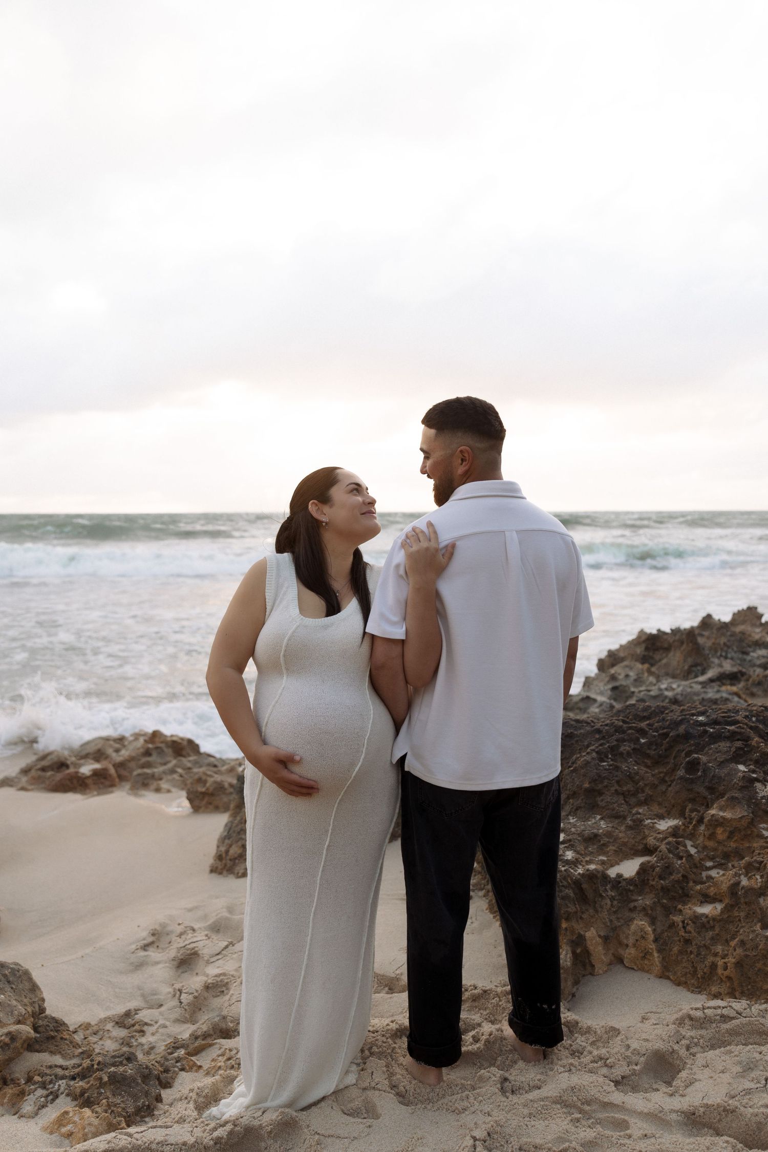 Pregnant woman in white maxi dress cradling baby bump on sandy beach at sunset.