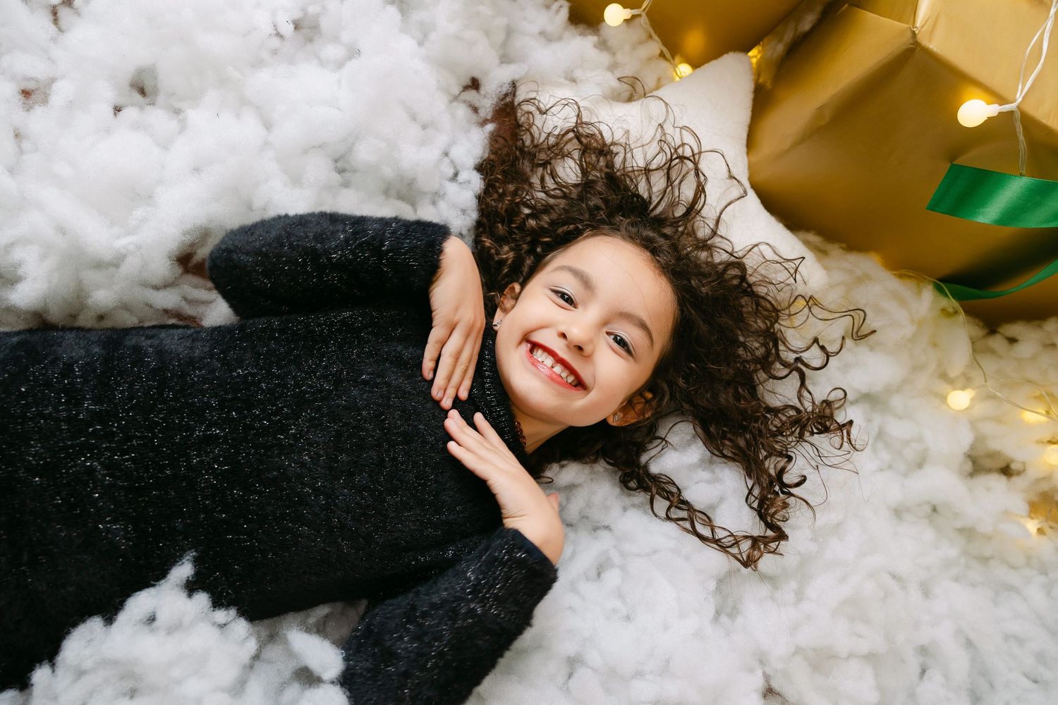Joyful young girl with curly hair laughing while lying on fluffy white cotton, wearing a dark knit sweater.