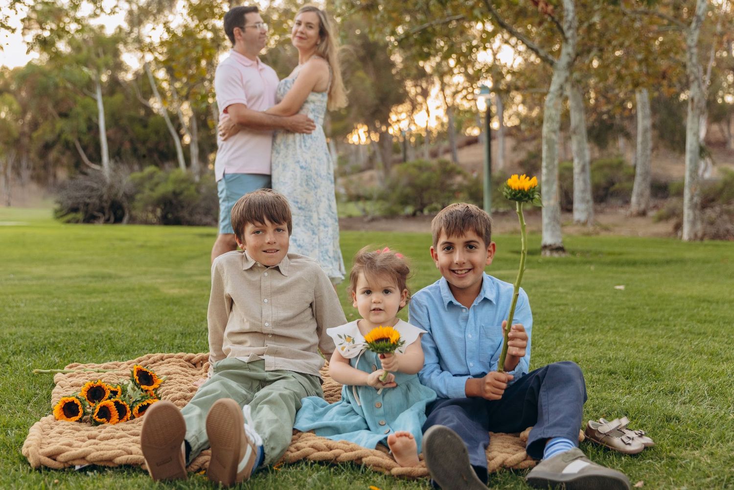 Family portrait with three boys holding sunflowers sitting on blanket while parents embrace in park.