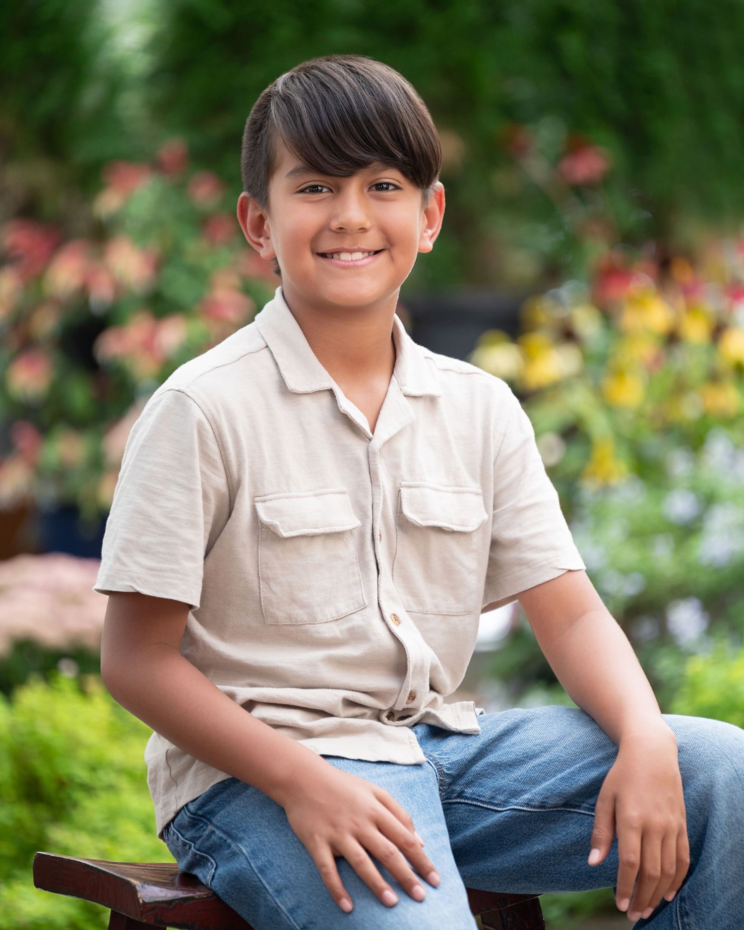 Smiling young boy in beige shirt and jeans sitting outdoors with colorful garden flowers in the background.