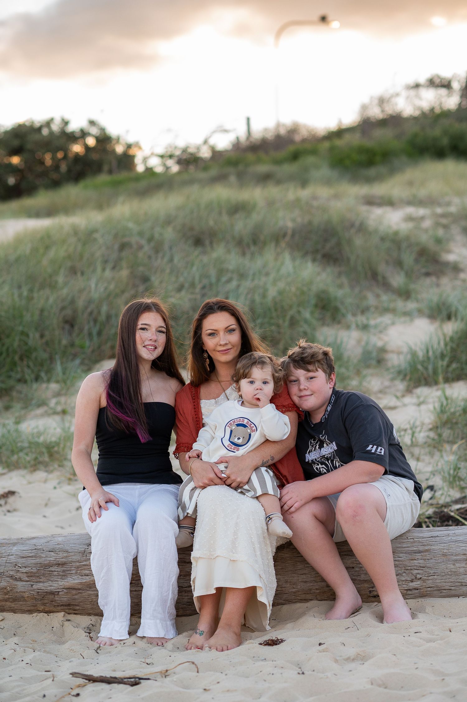 Three happy children sitting together on sandy beach dunes, smiling outdoors in summer attire.
