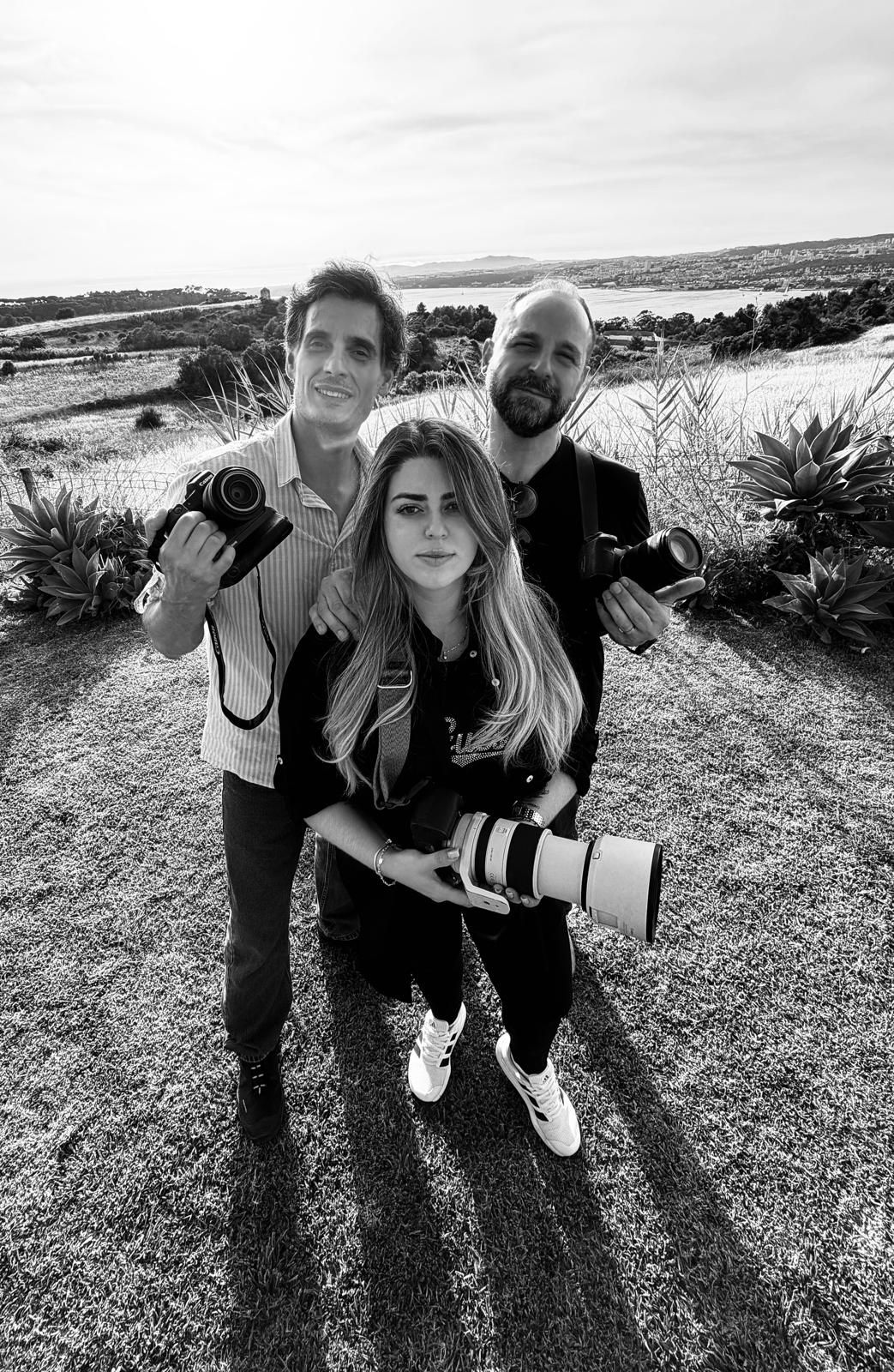 Three photographers posing outdoors in black and white, holding professional cameras with scenic landscape in background.