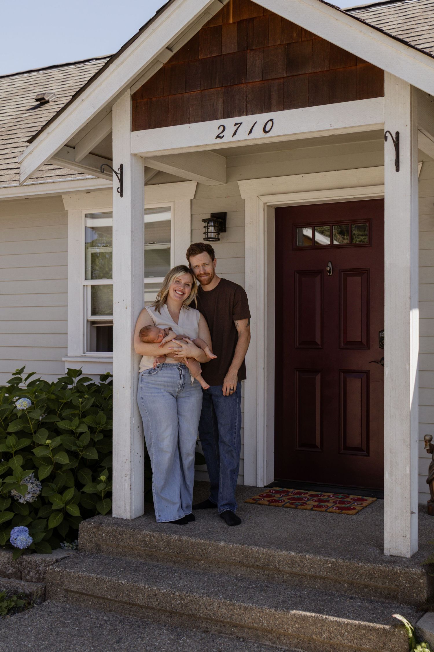 A young couple stands smiling on the front porch of their home at 2710, holding a newborn baby.