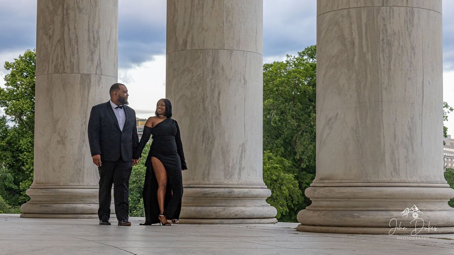 Couple in formal attire posing between grand white marble columns at a historic monument outdoors.