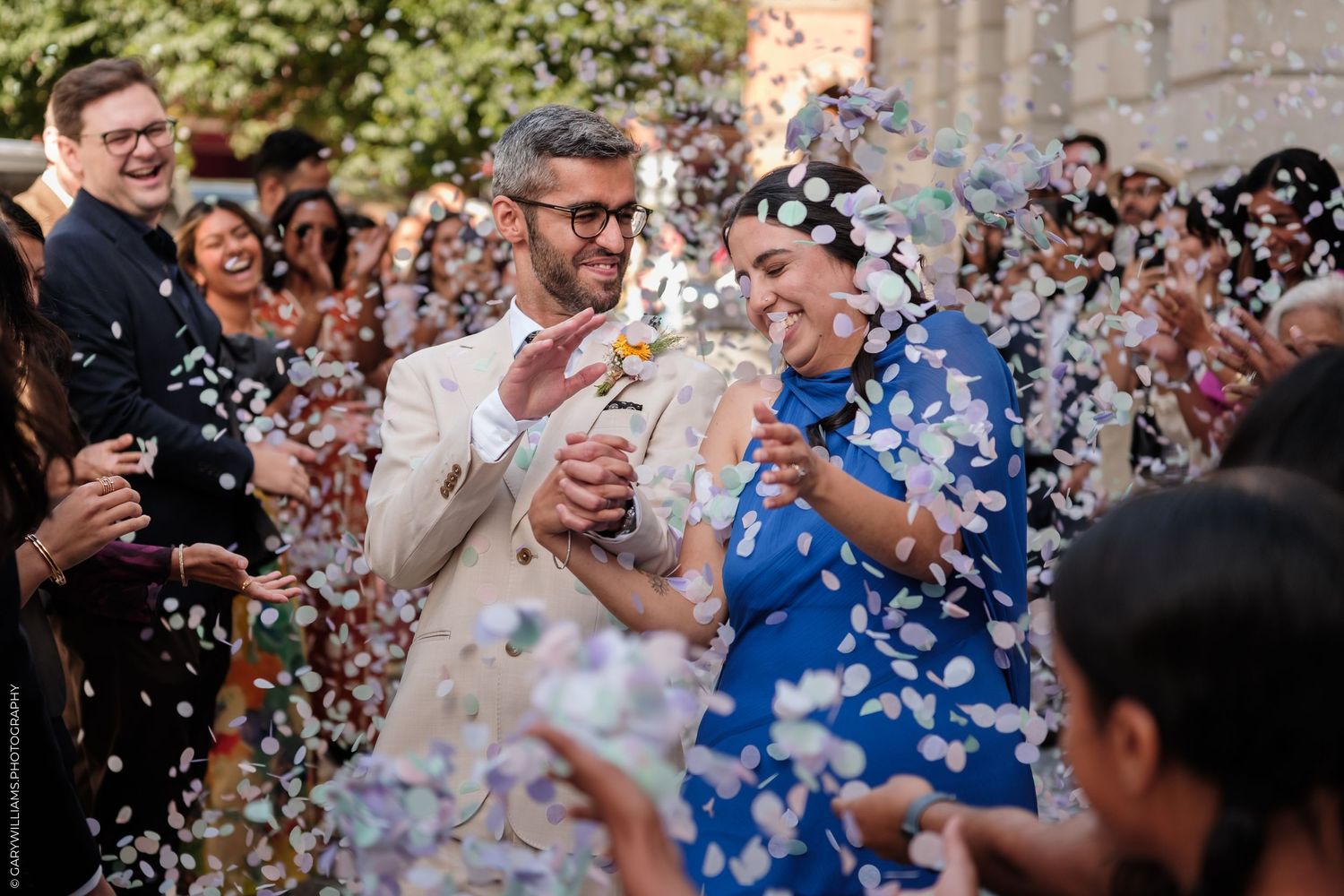Couple walks through confetti shower at outdoor wedding ceremony, guests cheering, bride in blue dress, groom in beige suit.