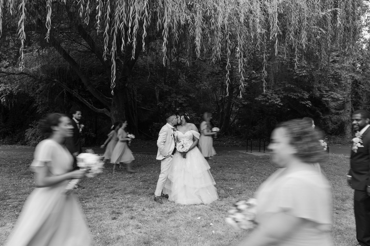 Black and white wedding photo of couple kissing under willow tree with blurred bridesmaids in foreground.