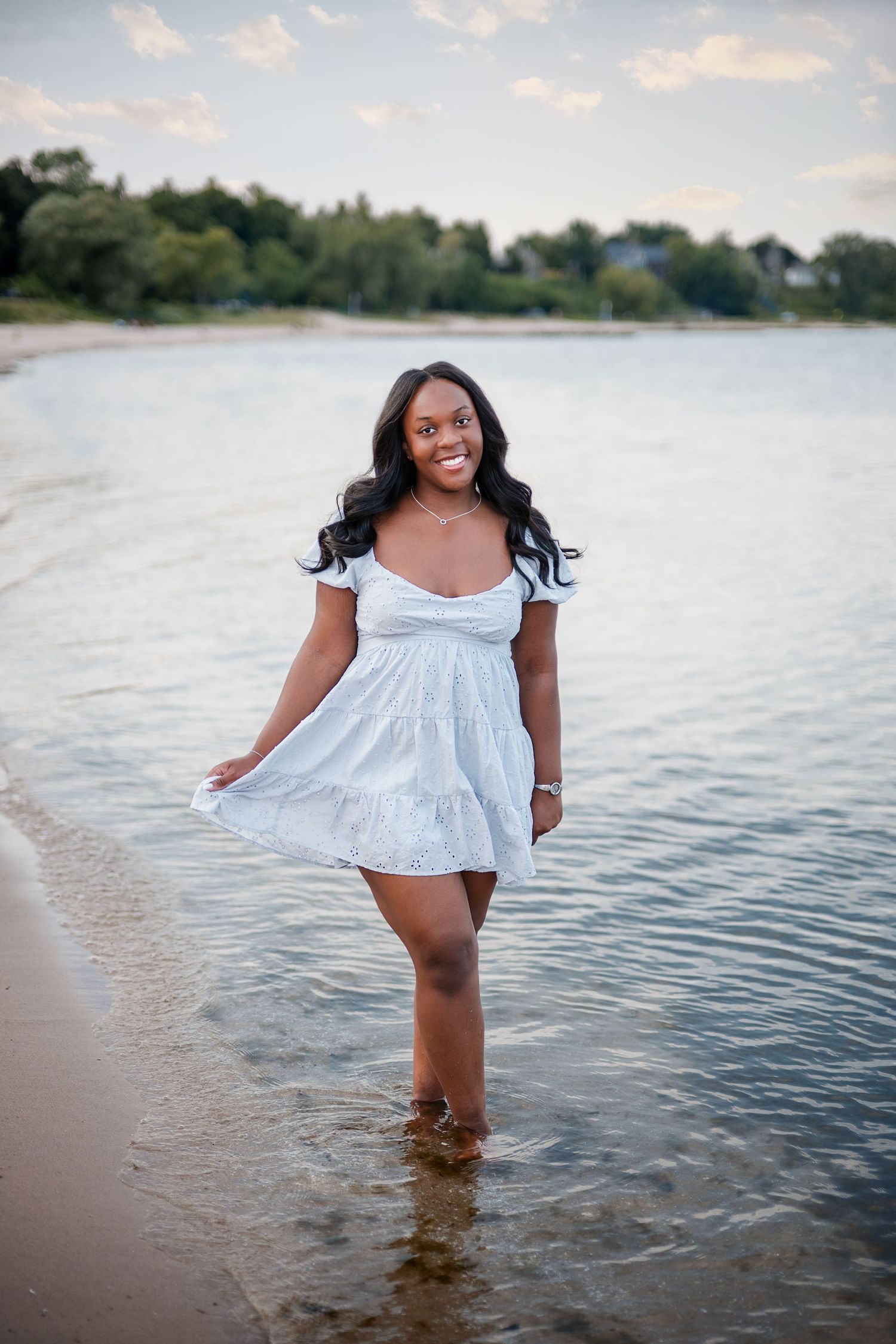 A smiling young woman in a white sundress stands in shallow beach water, posing confidently with trees in the background.
