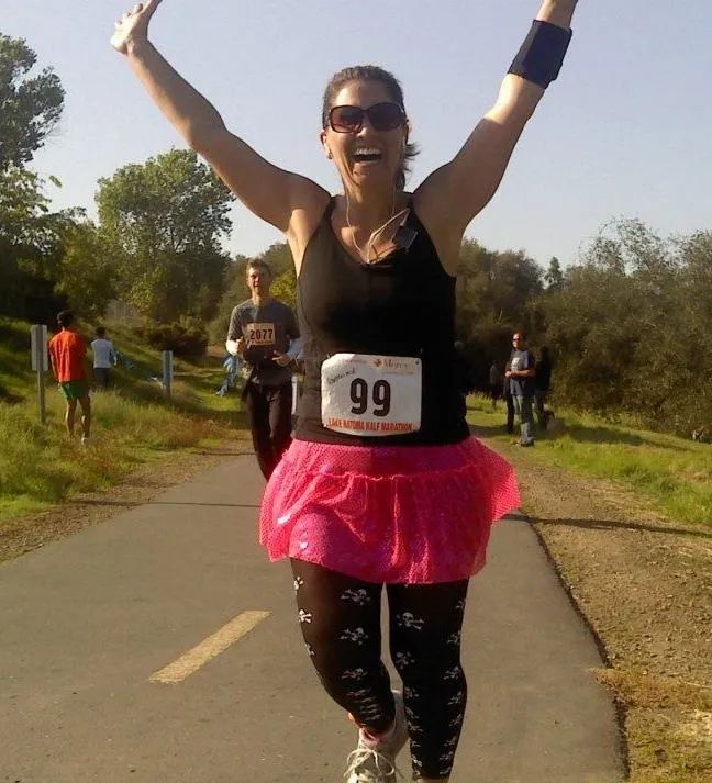 Woman in pink tutu and race bib number 99 celebrates with arms raised while running a 5K race on a paved path.