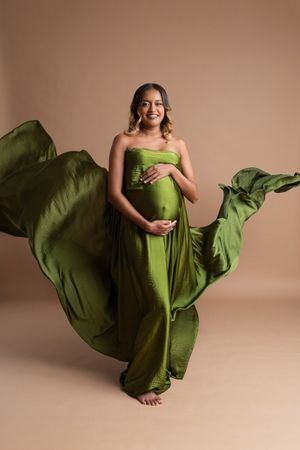 Pregnant woman in flowing olive green strapless gown with fabric billowing, smiling and cradling baby bump on beige background.