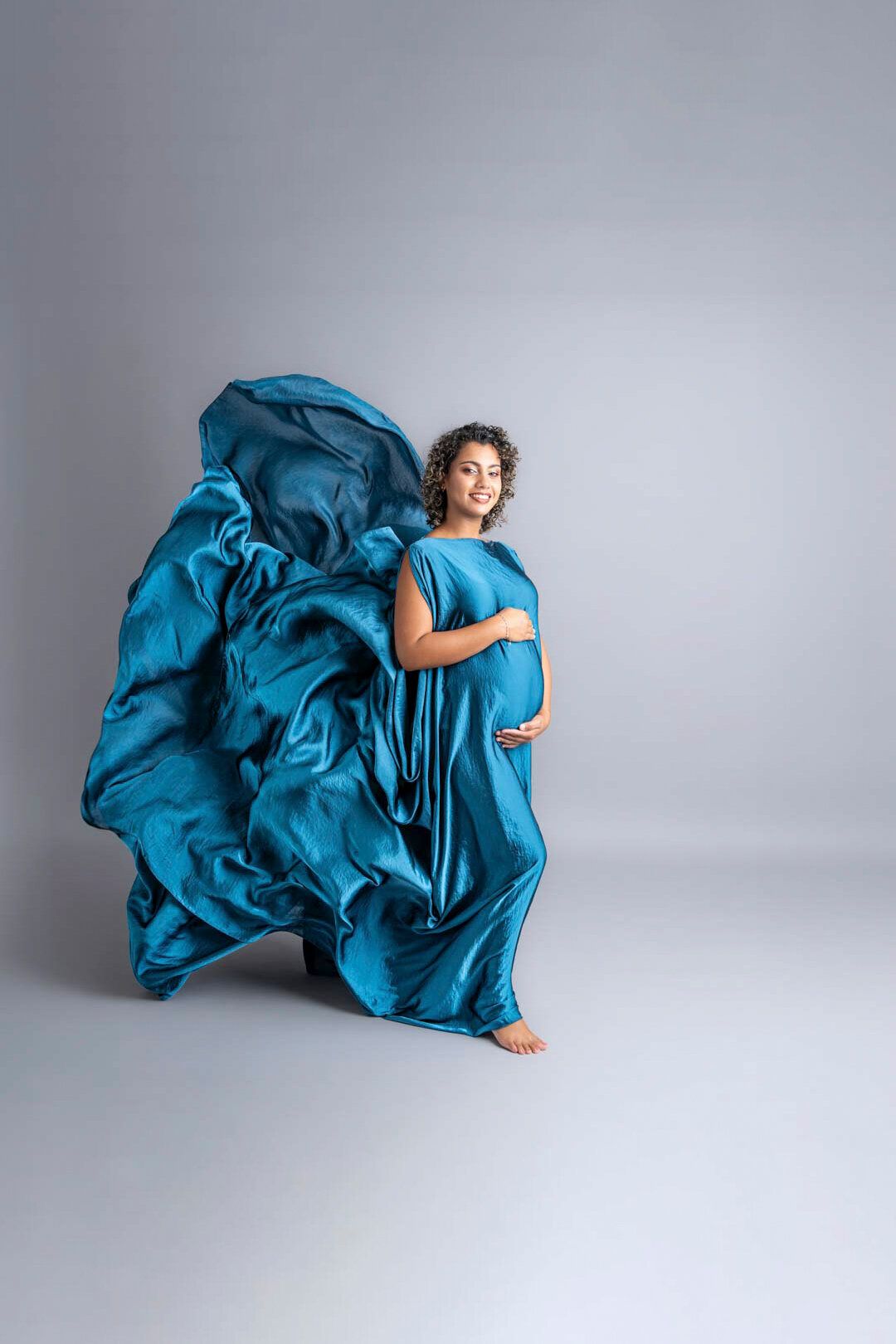 Pregnant woman in flowing teal silk gown with fabric billowing dramatically against gray studio background.