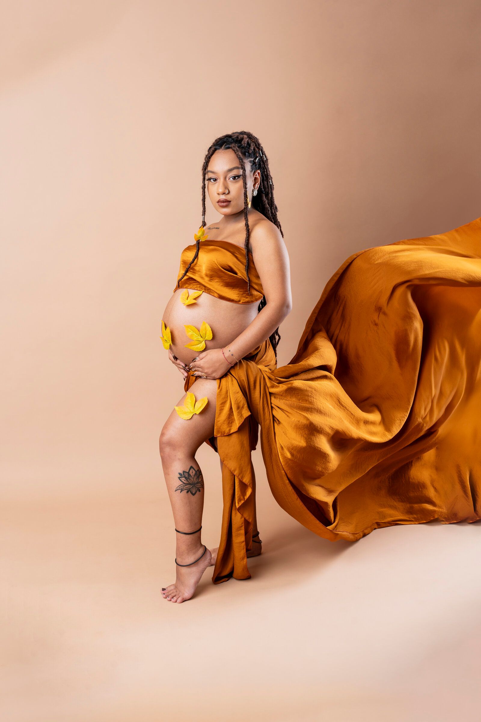 Pregnant woman in flowing golden satin fabric with yellow flowers, posing barefoot against beige background.