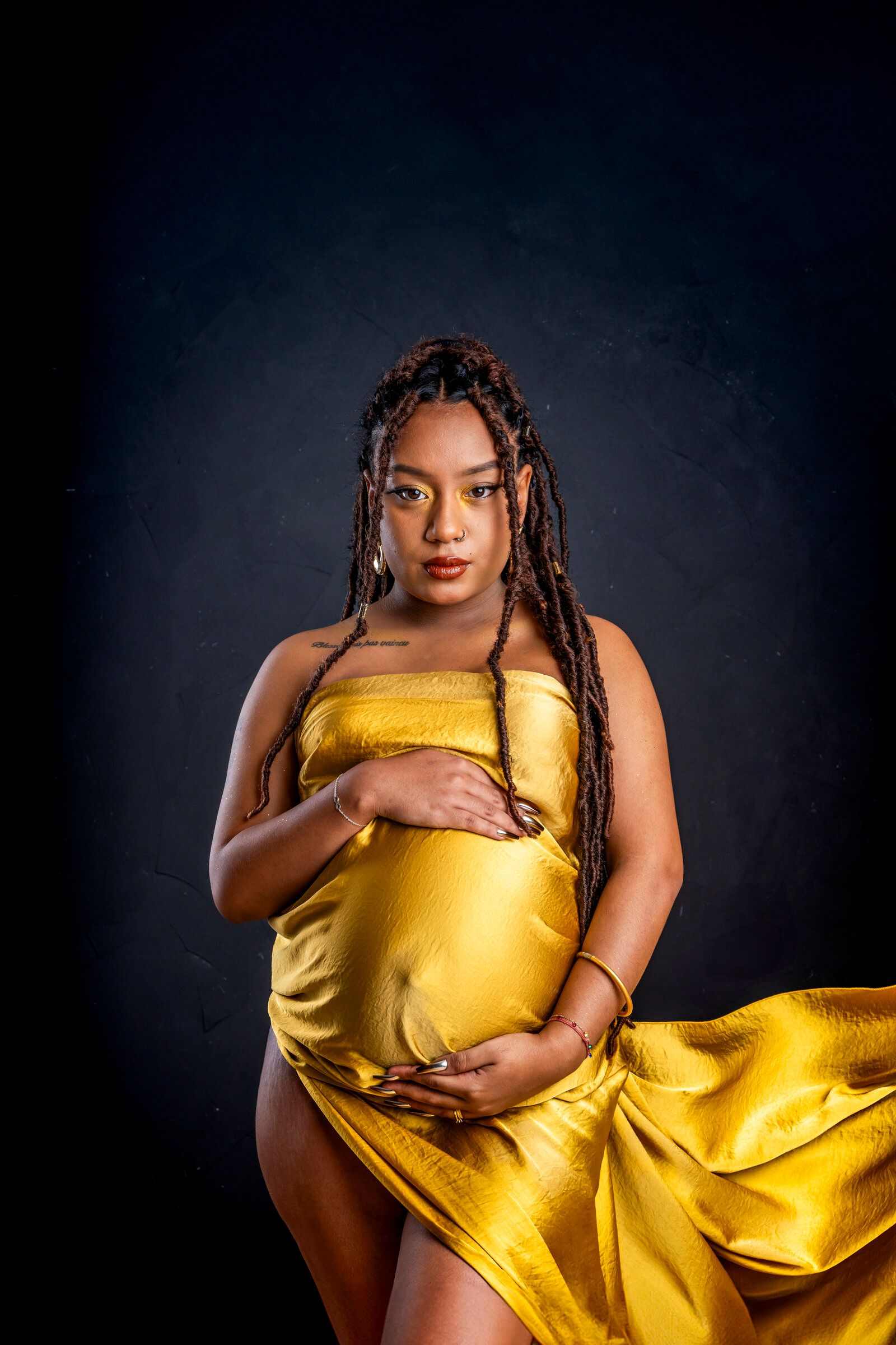 Pregnant woman in golden yellow satin dress cradling baby bump, wearing braids, posed against dark studio background.