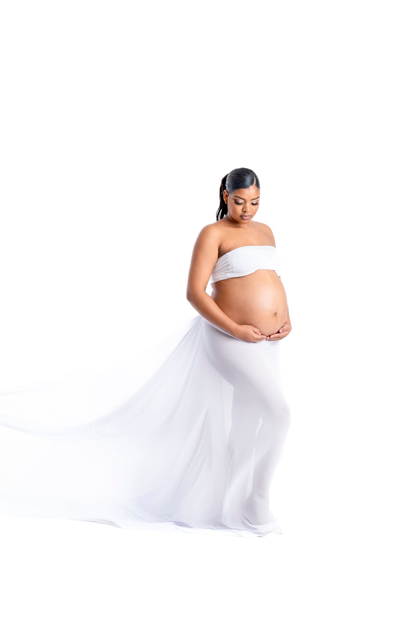Pregnant woman in white strapless bandeau and flowing sheer skirt cradling baby bump against white background.