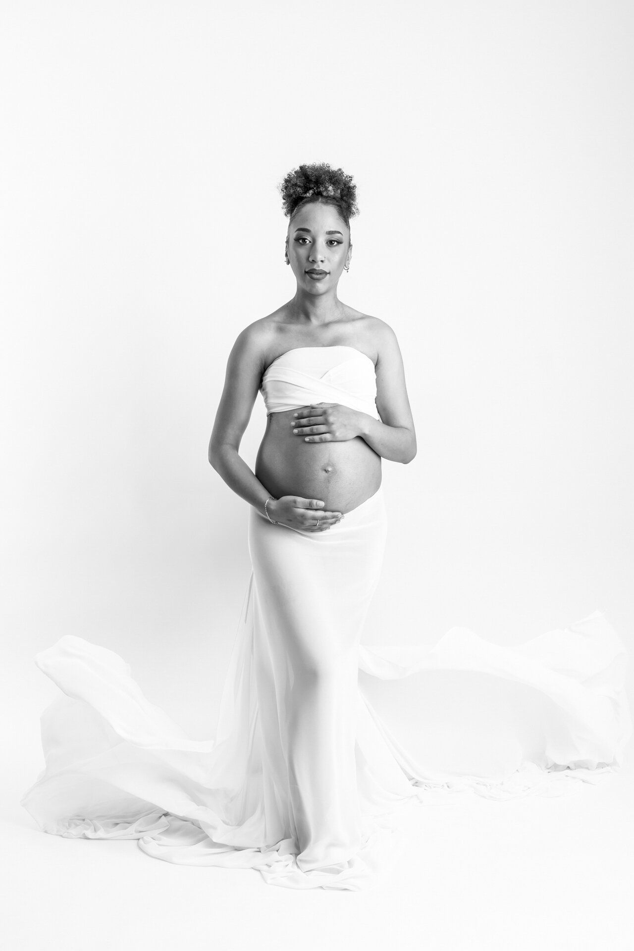 Black and white maternity photo of a pregnant woman in white draped fabric, cradling her baby bump in a studio setting.