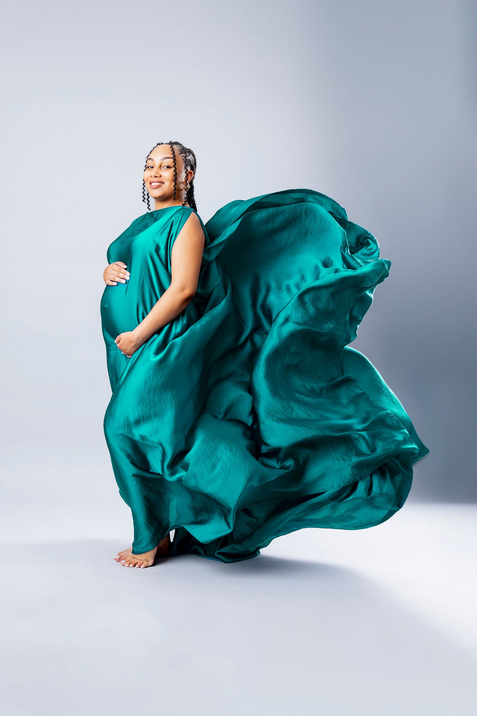 Pregnant woman in flowing teal silk dress, smiling gracefully against a light gray studio background.