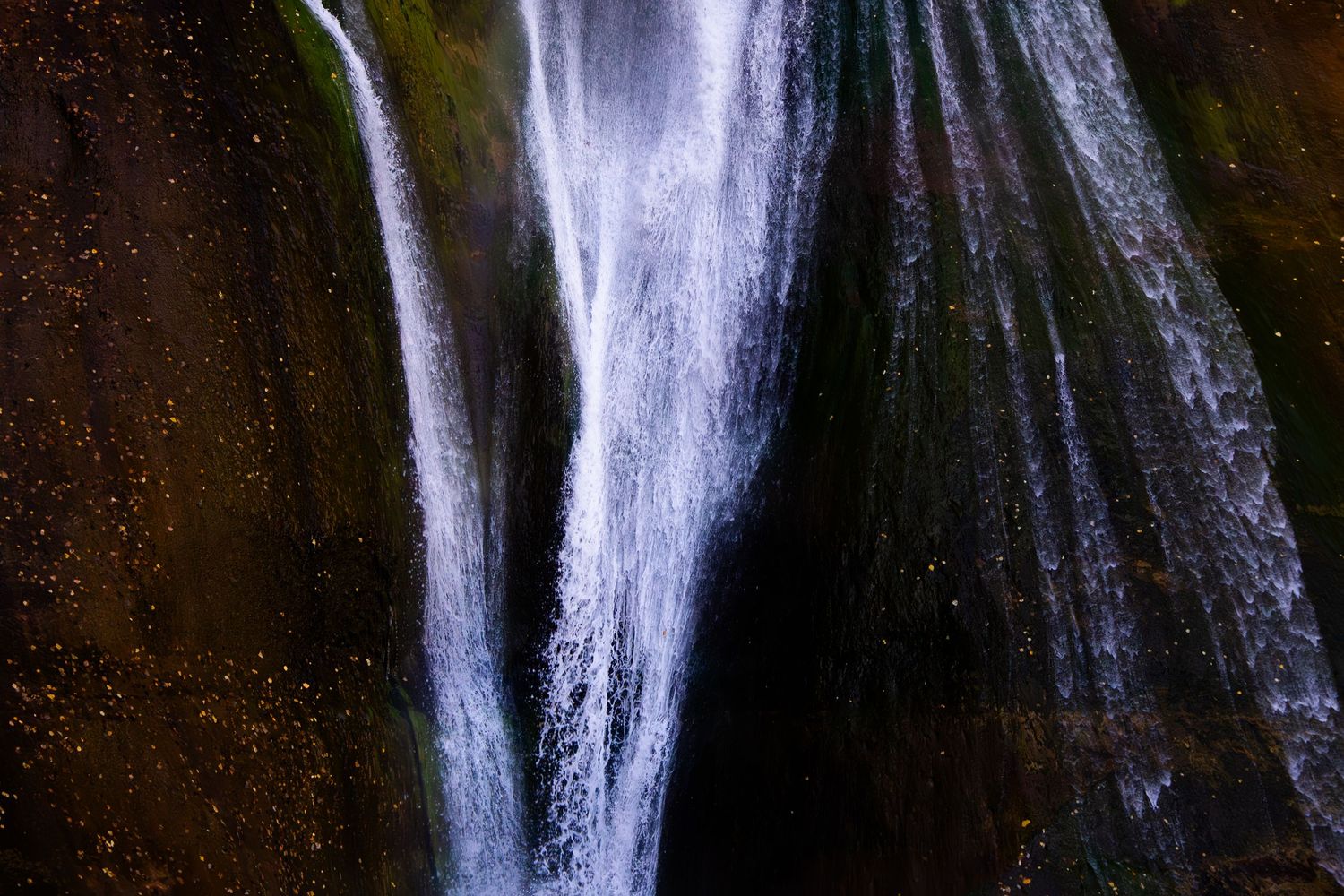 Close-up of cascading waterfalls flowing over dark rocky cliffs with lush green moss in a natural setting.