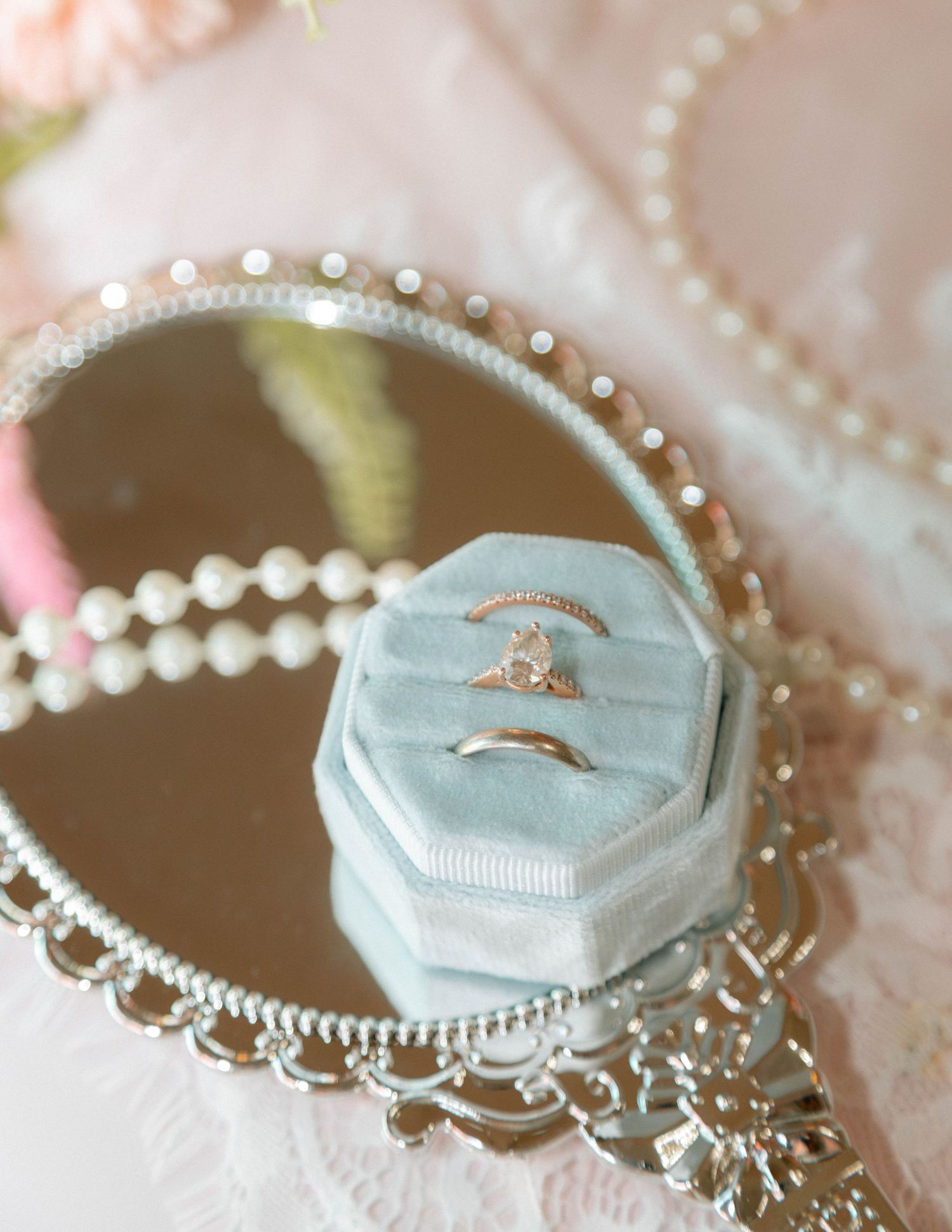 Light blue velvet octagonal ring box with wedding bands on a silver mirrored tray with pearl necklace.