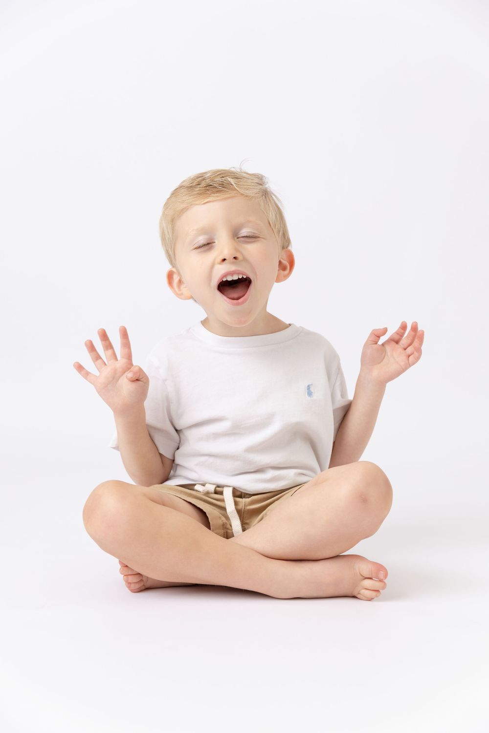 Young blonde boy sitting cross-legged, eyes closed, mouth open, hands raised, wearing white t-shirt and tan shorts on white background.