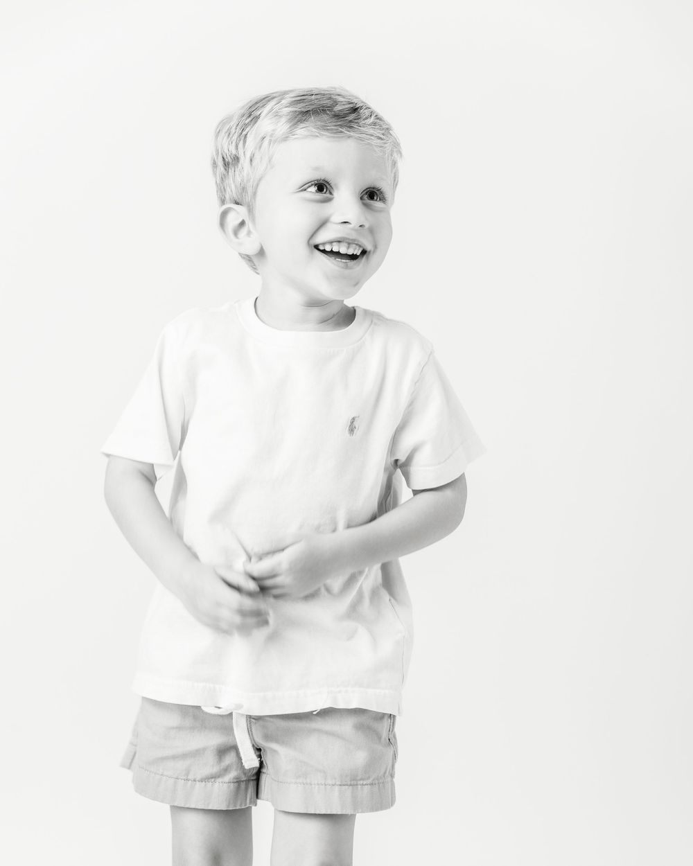 Smiling young boy in white t-shirt and khaki shorts poses cheerfully in a black and white studio portrait.
