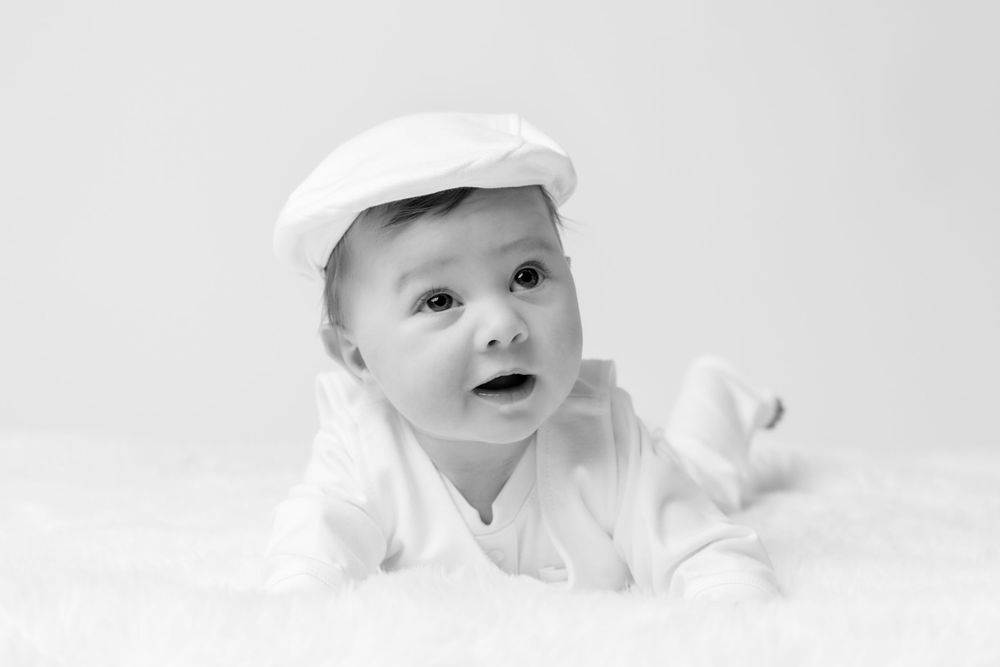 Black and white photo of an adorable baby in a white cap and outfit lying on a fluffy white surface.