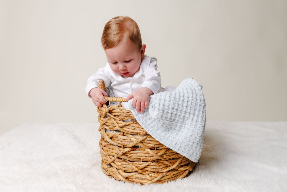 Adorable baby sitting inside a wicker basket with a gray knit blanket, wearing a white outfit.