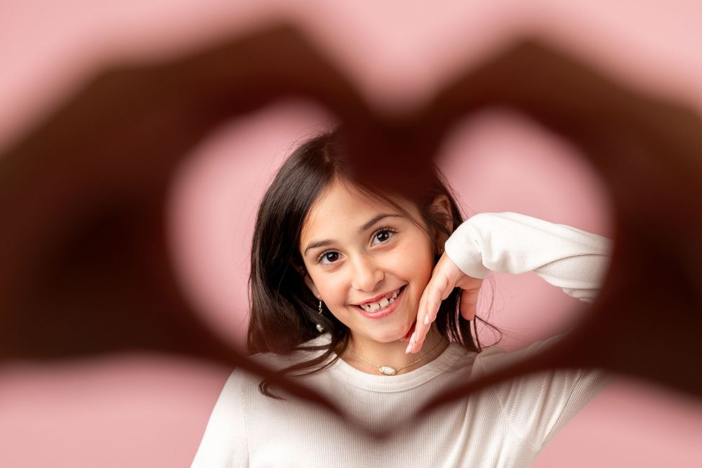 A smiling girl framed by hands forming a heart shape against a pink background.