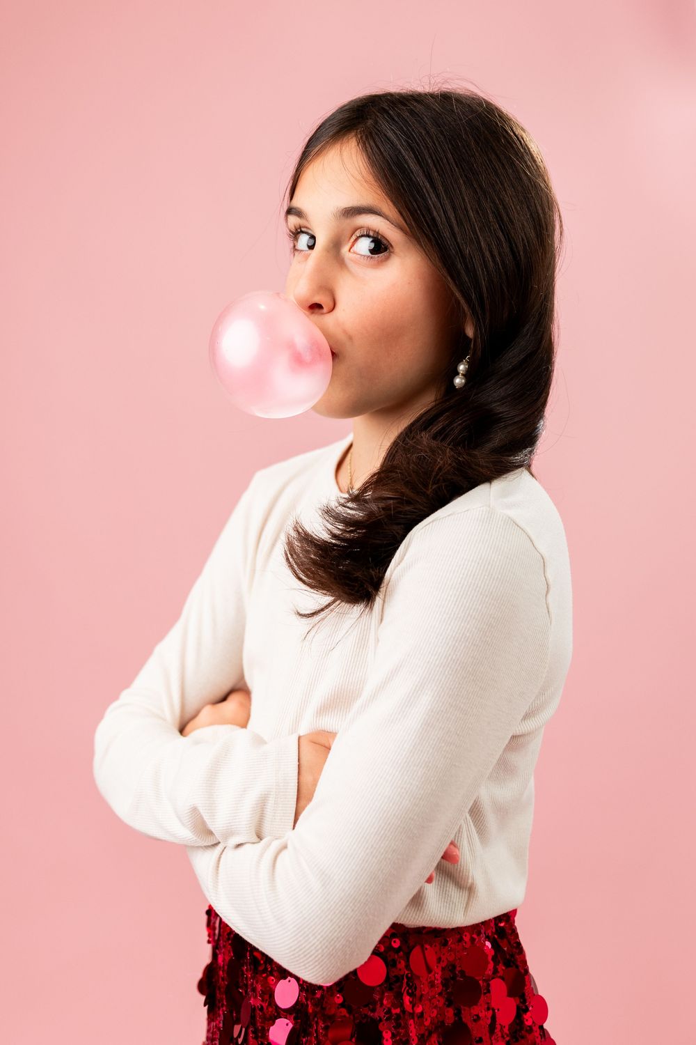 Young woman with dark hair blowing a pink bubble gum bubble, arms crossed, wearing white top and red skirt.
