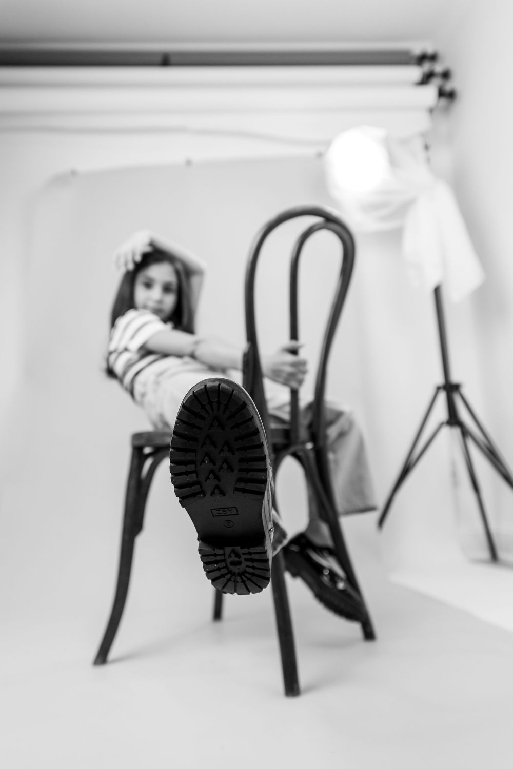 Black and white studio photo of girl on chair with boot sole in foreground, lighting equipment visible.