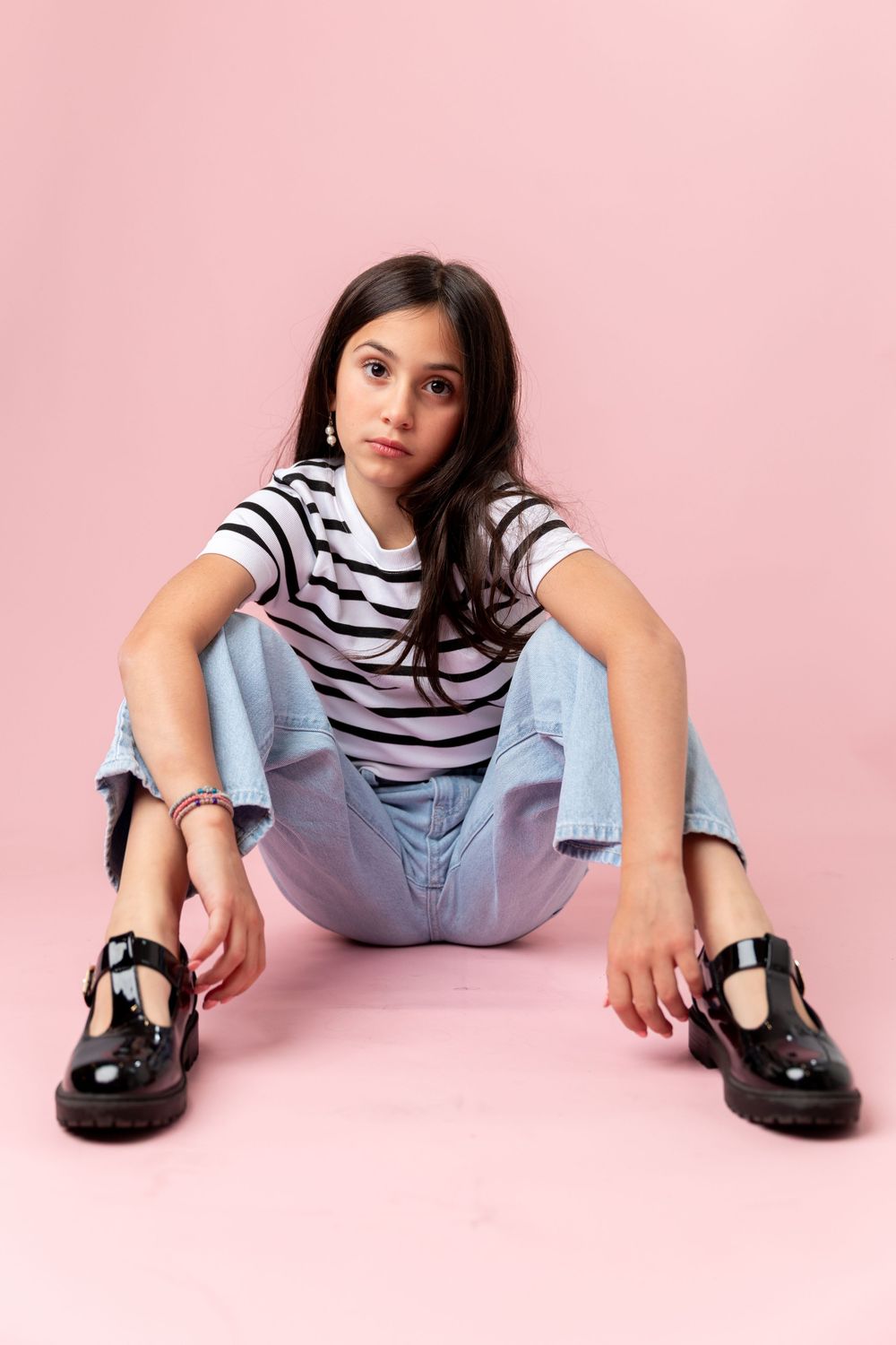 Young girl in striped tee, jeans, and black patent shoes sitting on pink background.