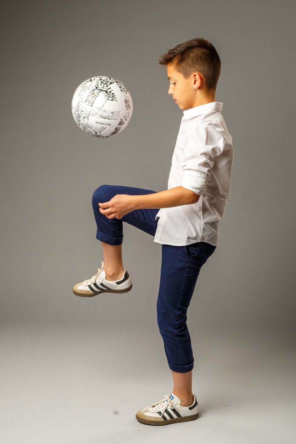 Young boy in white shirt and blue pants juggling a soccer ball on his knee against a gray background.