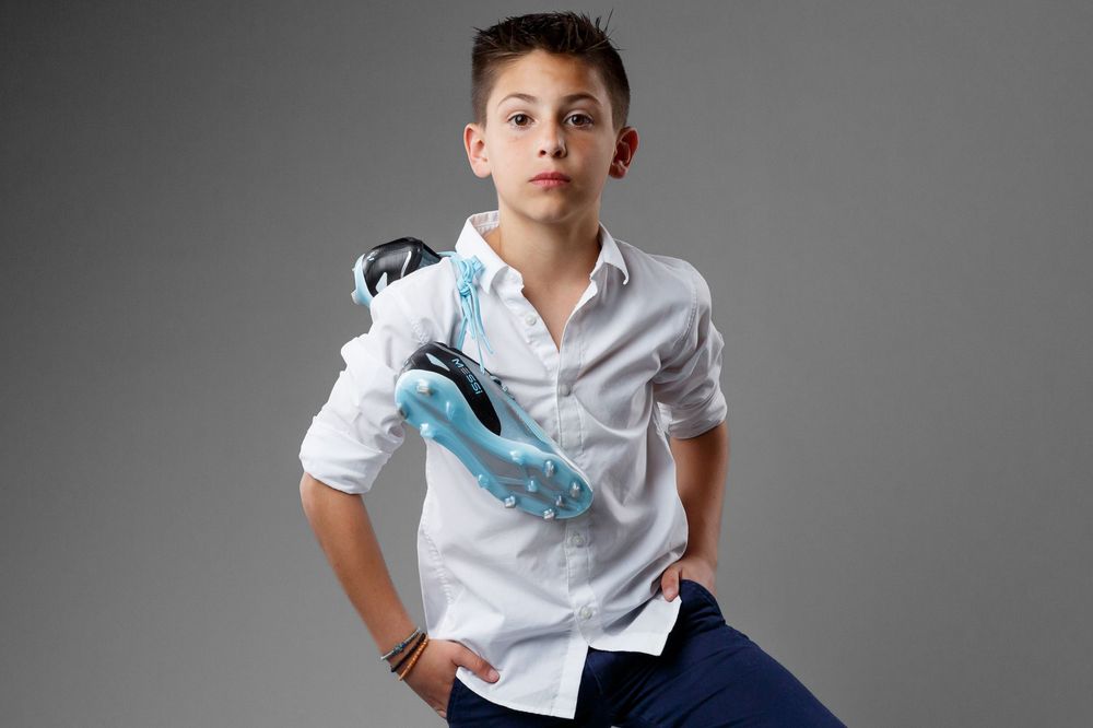 Young boy in white shirt holding soccer cleats over his shoulder against a gray background.