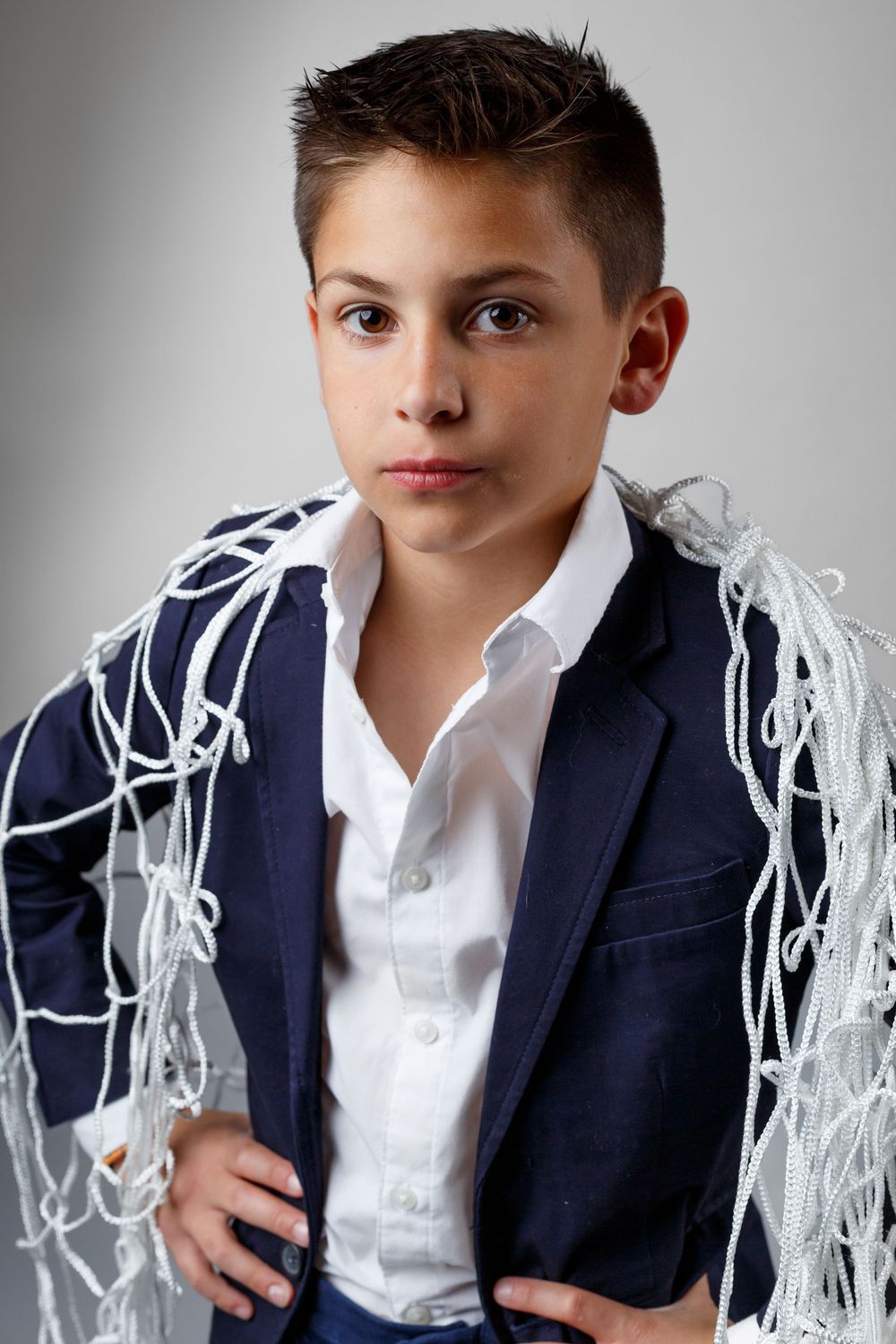 A young boy in a navy blazer and white shirt draped with a white rope net, posing confidently.