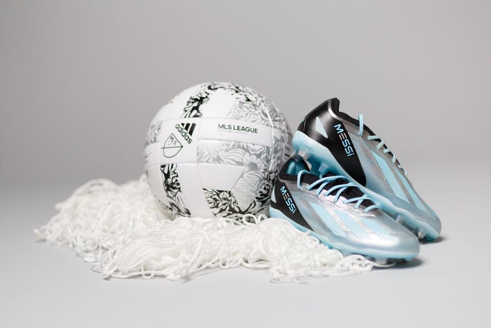 Silver and teal soccer cleat beside a white MLS League ball resting on a white net against a gray background.