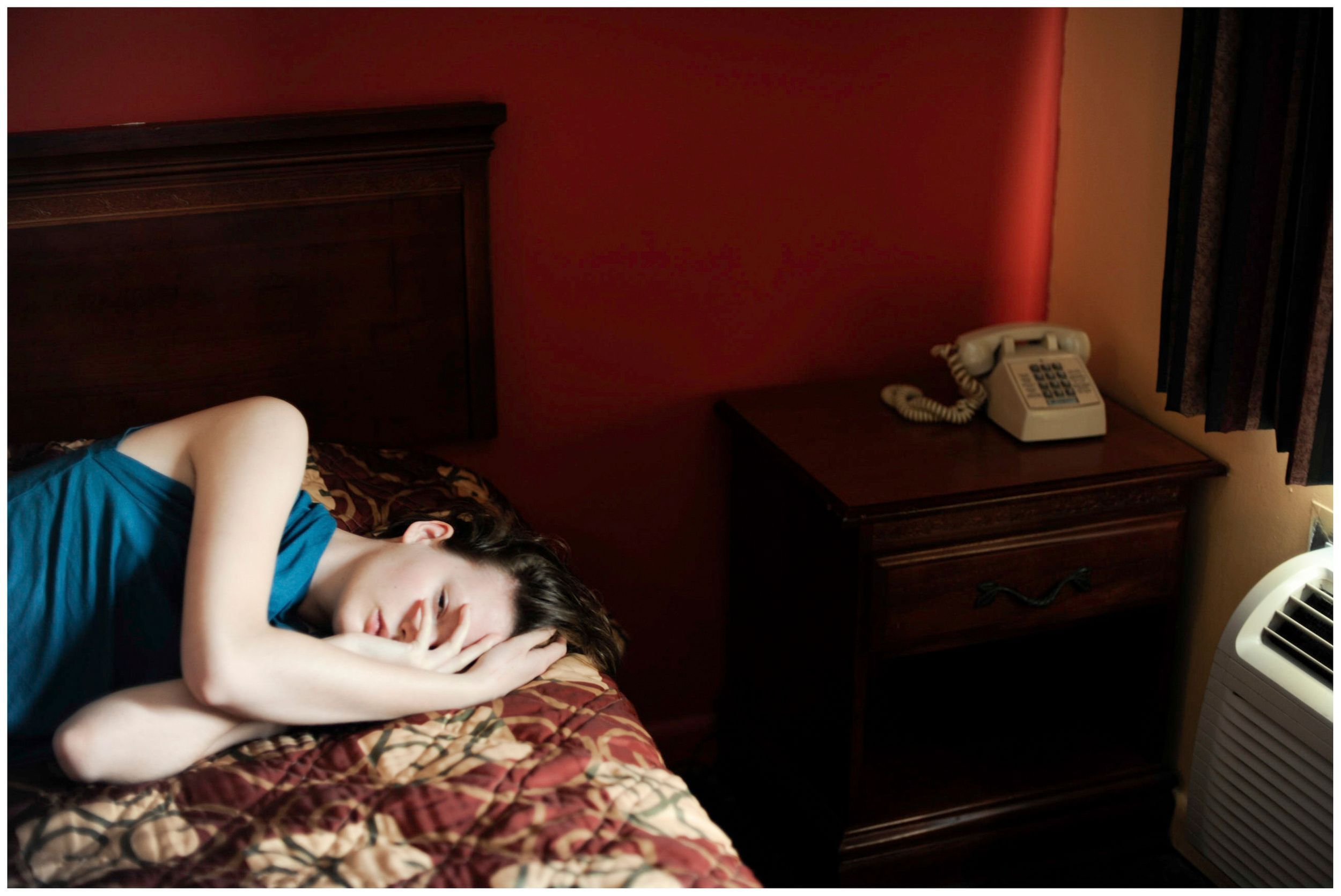 Woman in blue dress sleeping on a floral bedspread in a dimly lit motel room with red walls and a nightstand.