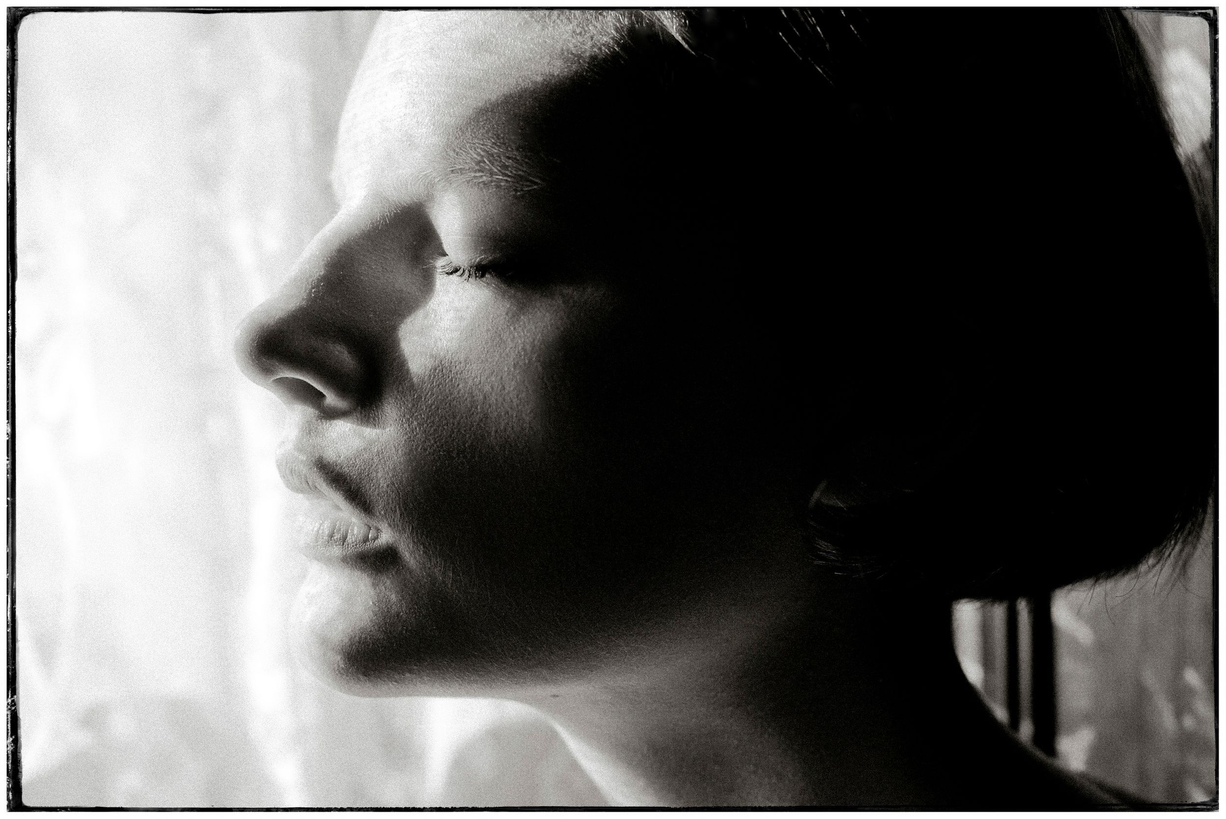 Black and white profile portrait of a woman with closed eyes, soft light illuminating her face near a window.