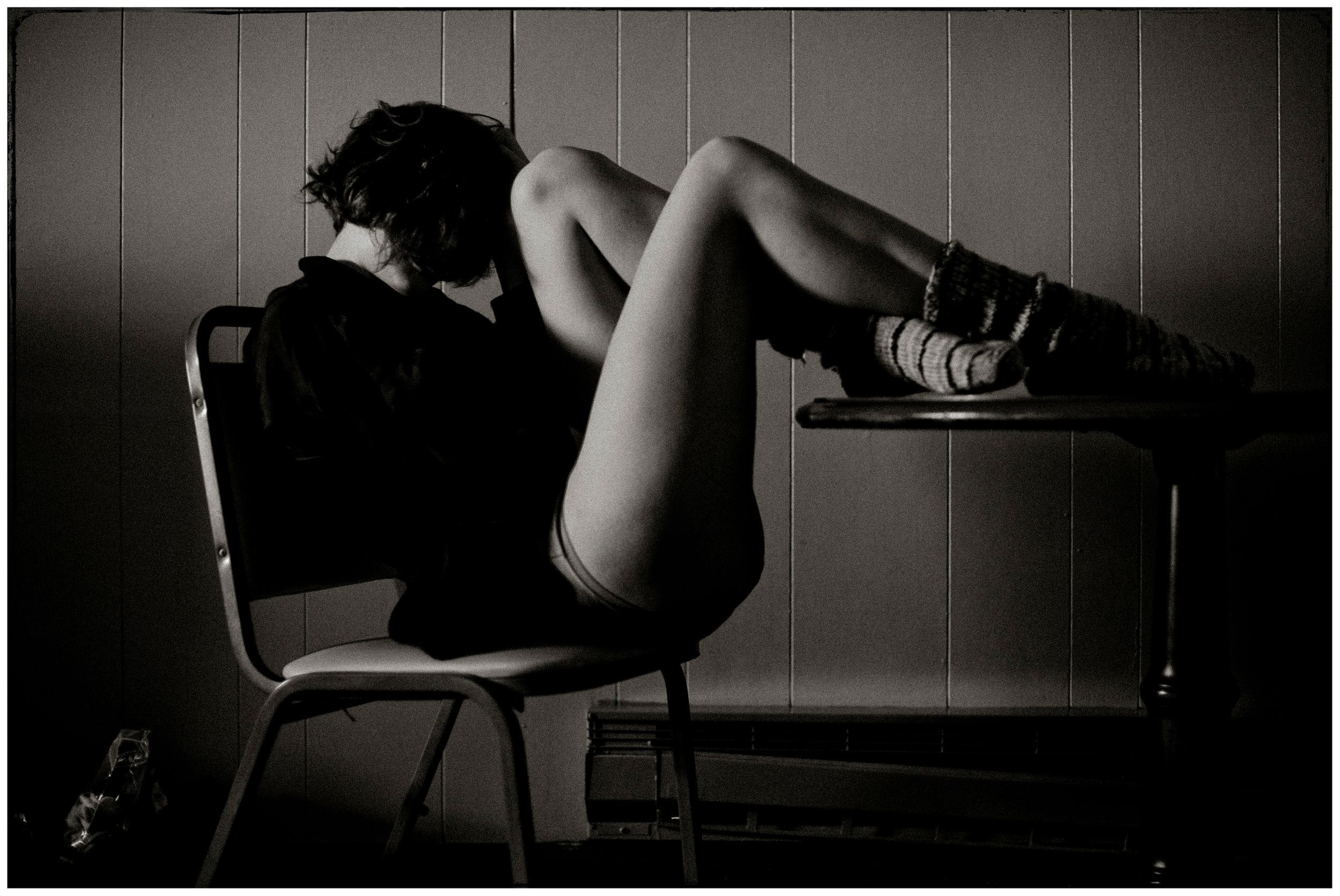 Black and white photo of a woman reclining on a chair with legs on a desk in a dimly lit room.
