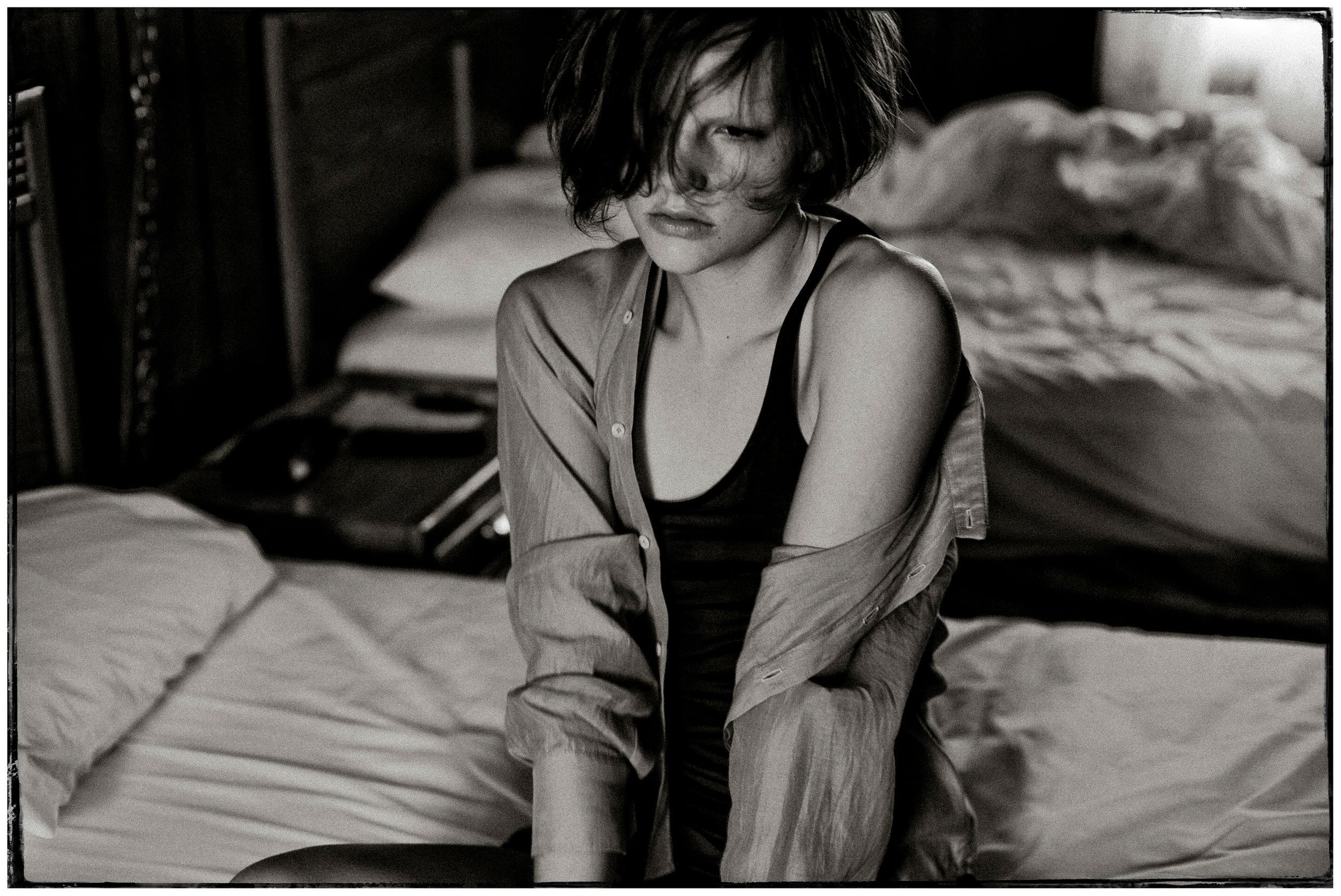 Black and white photo of a young woman sitting on a bed, wearing a tank top and open shirt, looking down.