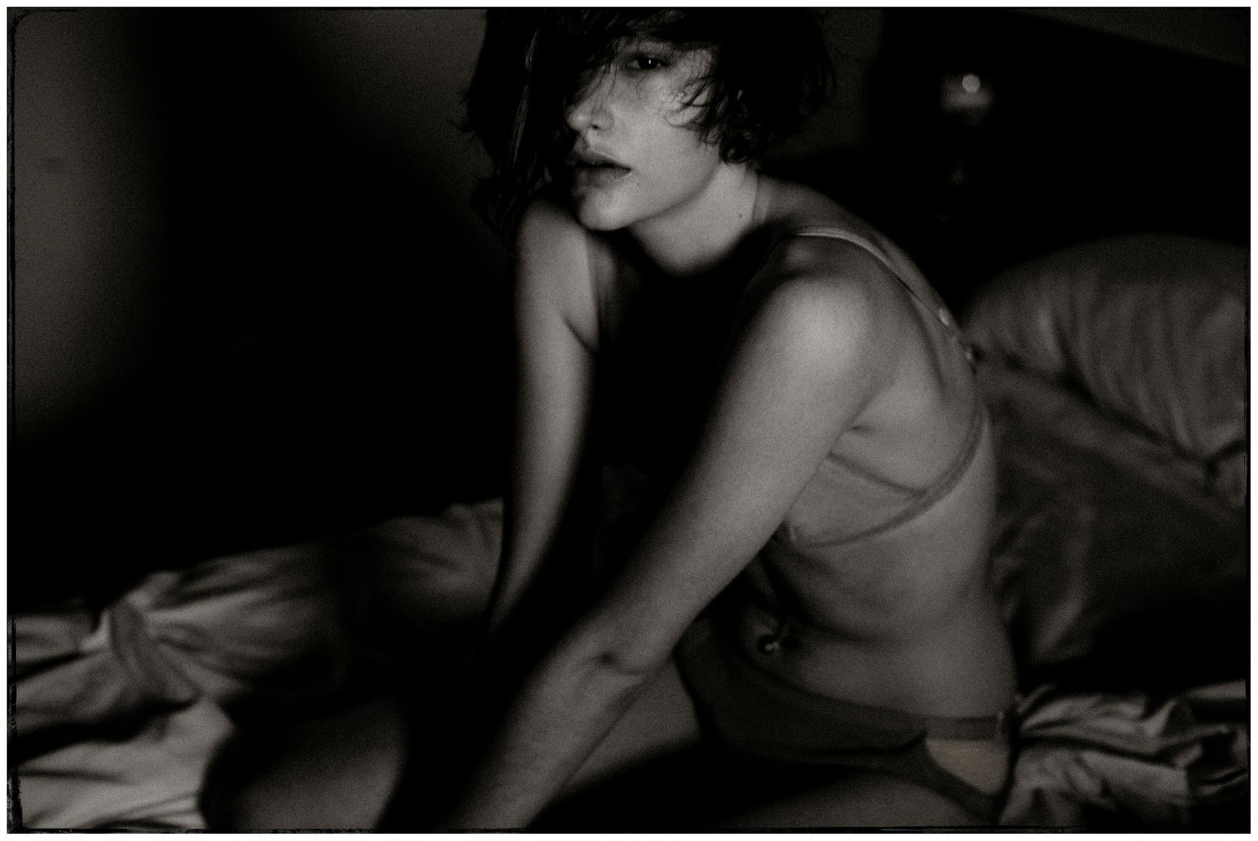 Black and white photo of a woman in lingerie sitting on a bed in moody dramatic lighting.