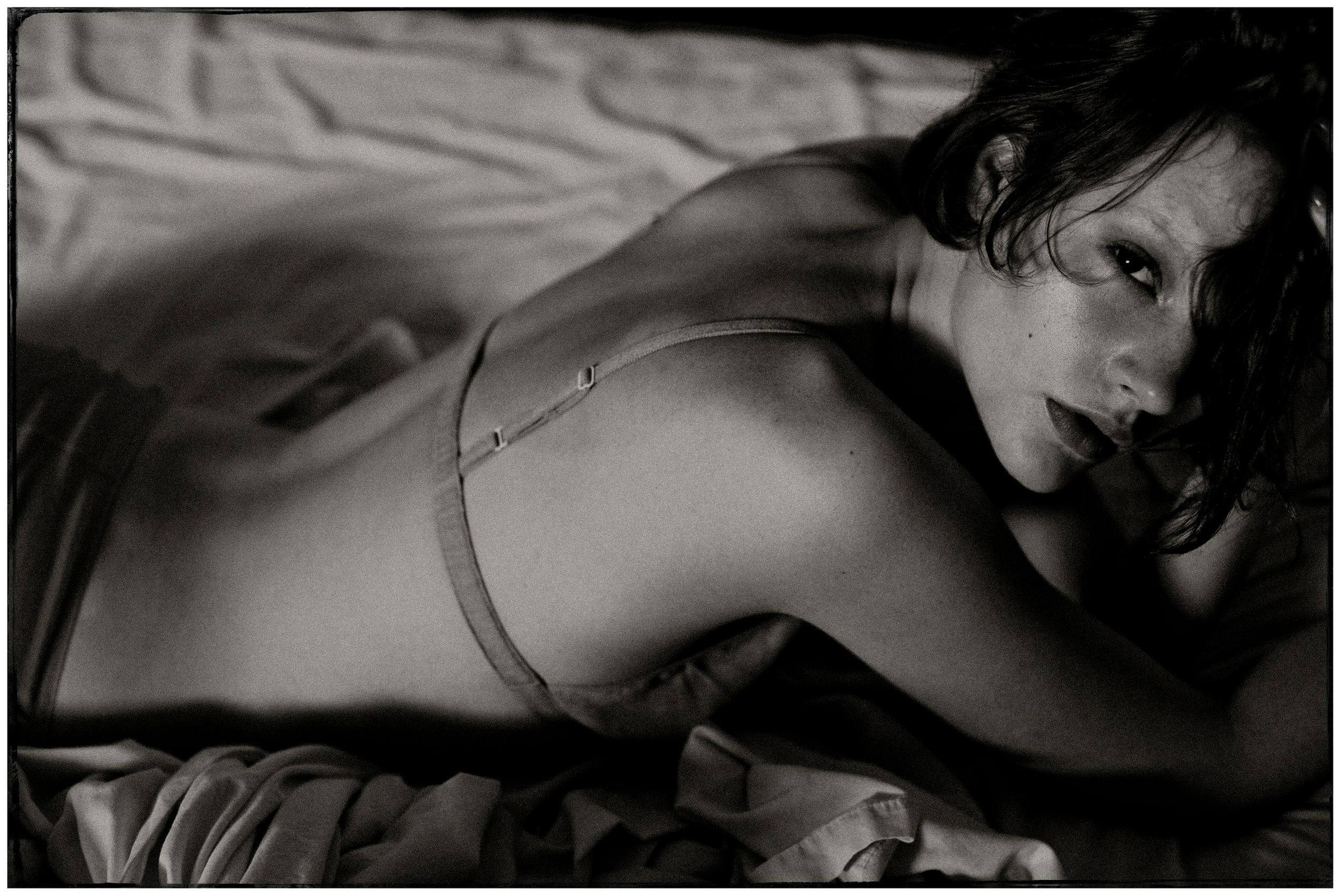 Black and white photo of a woman lying on a bed wearing a bra, looking over her shoulder.