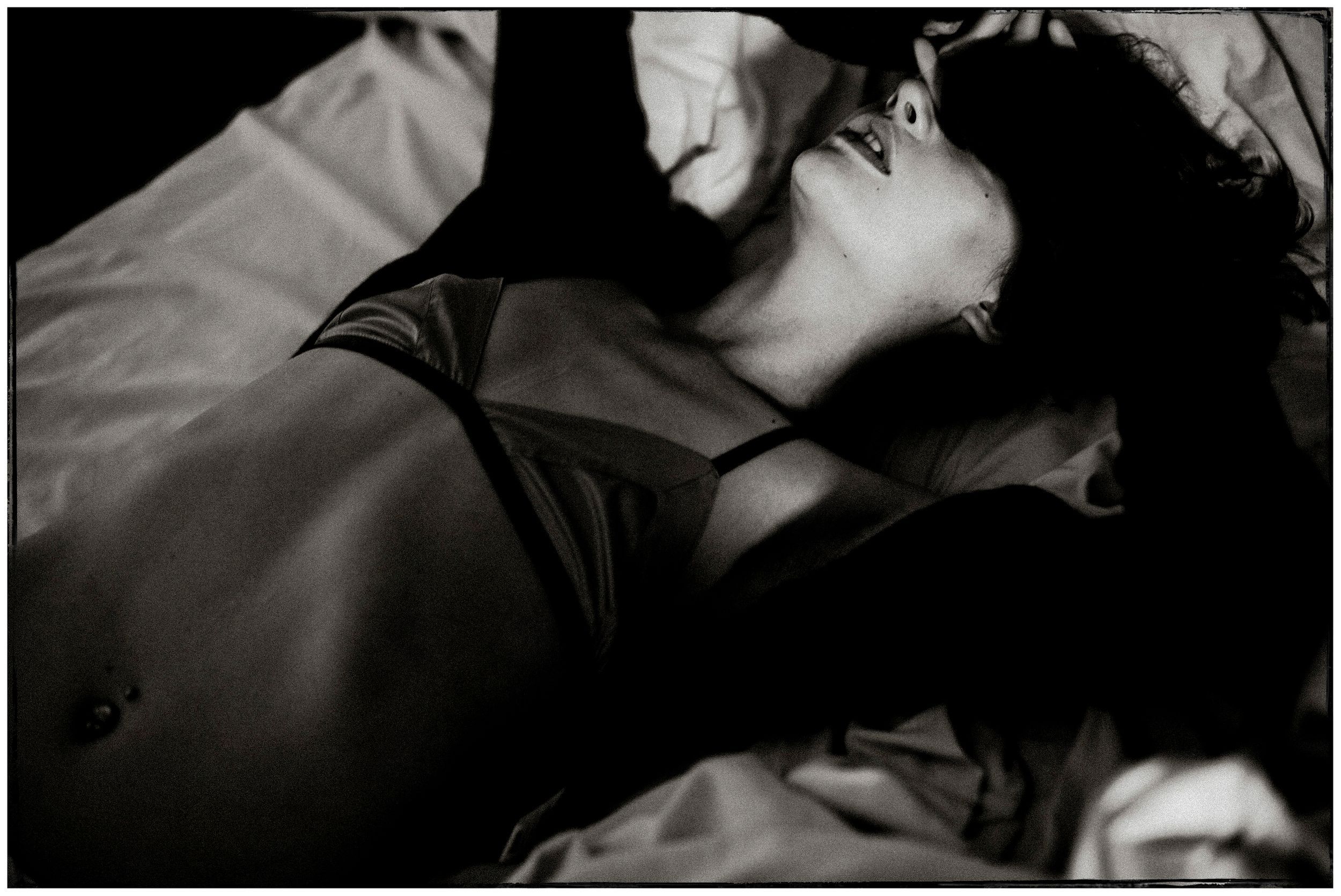 Black and white photo of a woman in lingerie lying on white bedding in dramatic light.