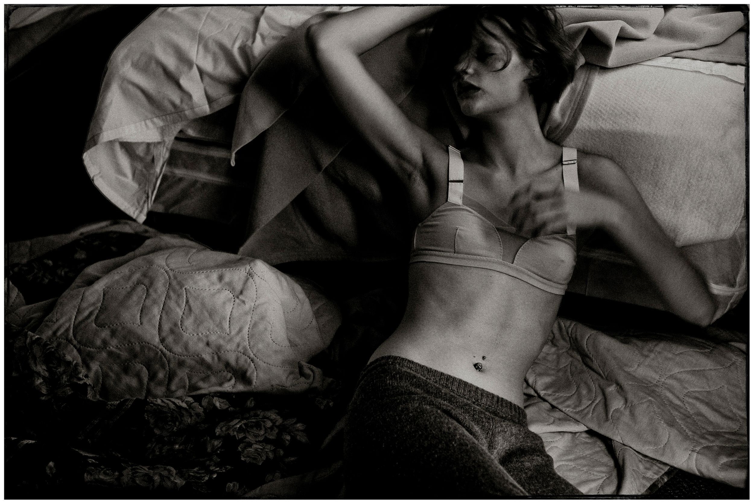 Black and white photo of a woman in a bra lying on rumpled bed sheets in a relaxed pose.