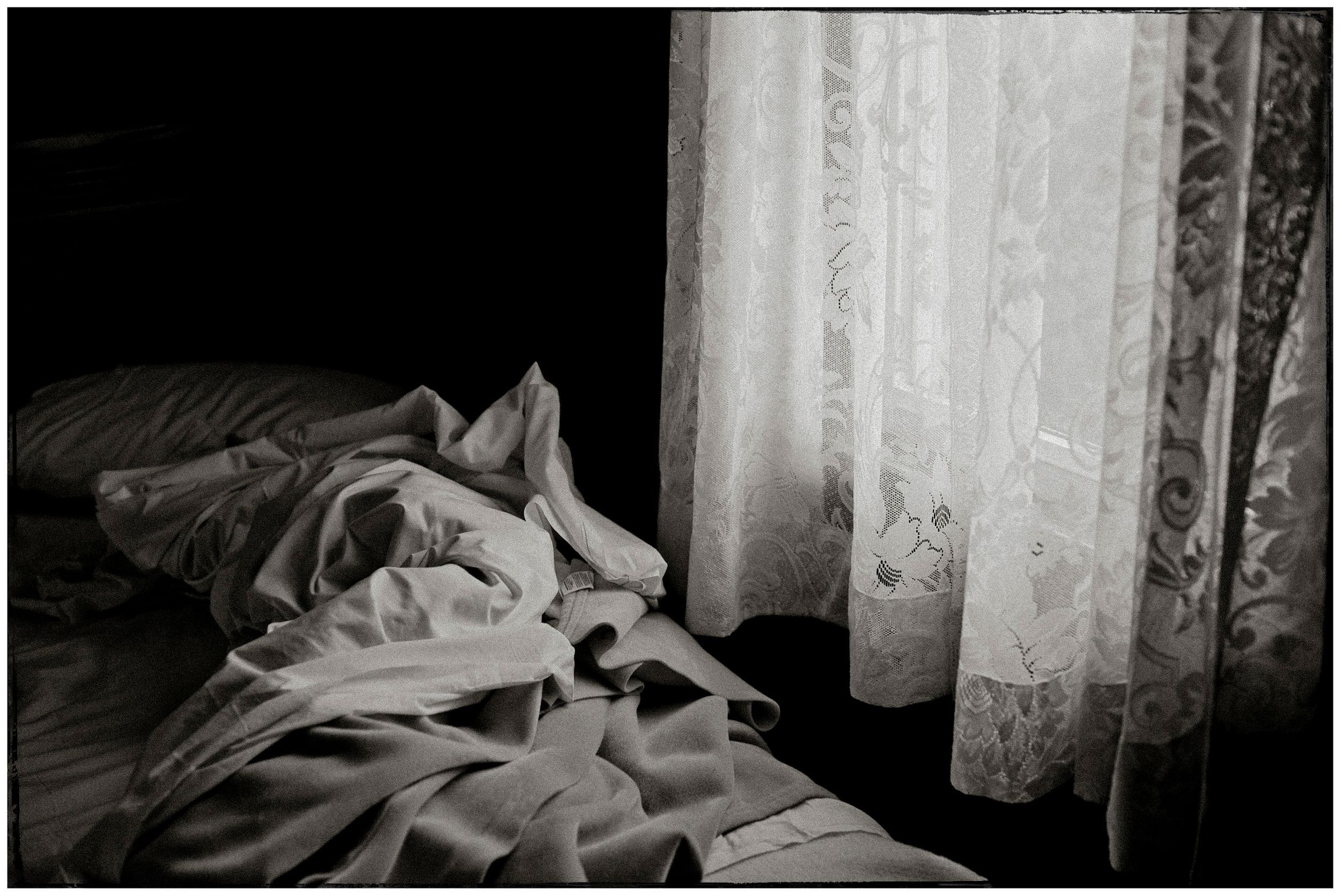 Black and white photo of rumpled bed sheets beside sheer lace curtains with soft light filtering through.