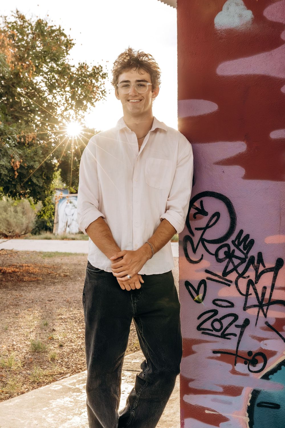 Smiling young man in white shirt and black jeans leaning against graffiti pillar in sunlight.