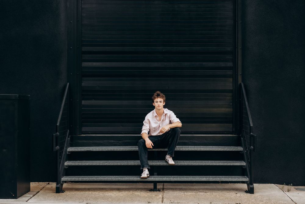 Young person sitting on dark metal stairs against a black wall, wearing casual white shirt and jeans.