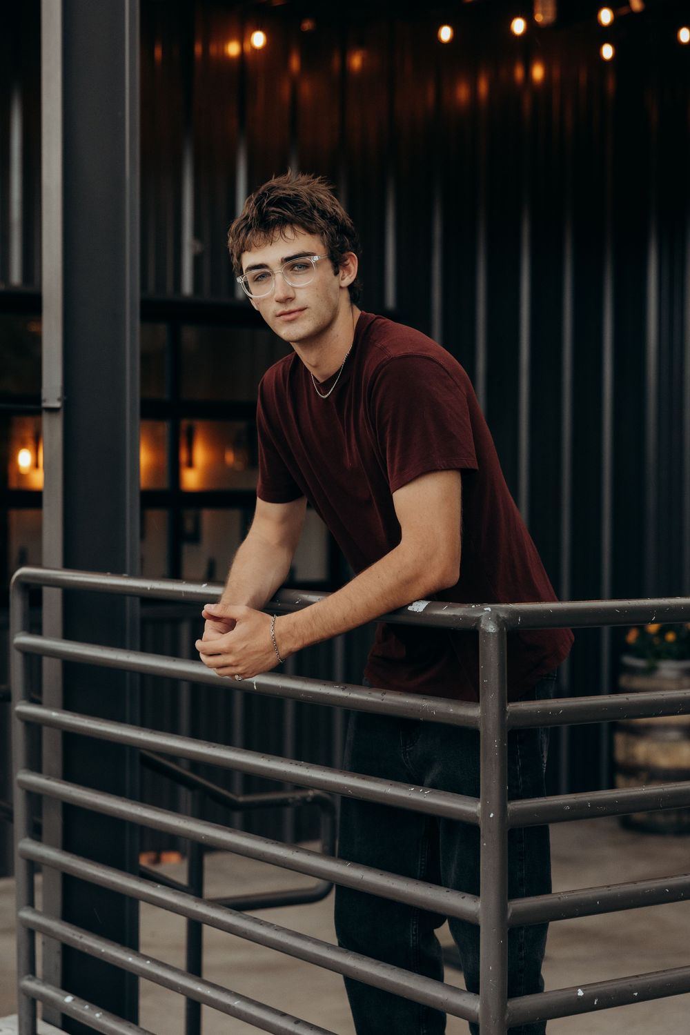 Young man in maroon t-shirt and glasses leaning on metal railing with bokeh lights in background.